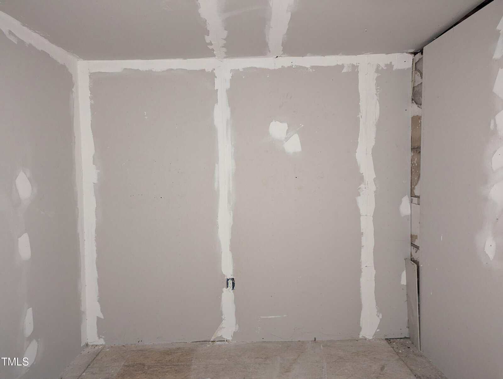 A photograph of an unfinished room with drywall that has been installed and taped but not yet painted. The floor appears to be concrete, and there are no fixtures or decorations in the room. The walls show signs of recent construction work.