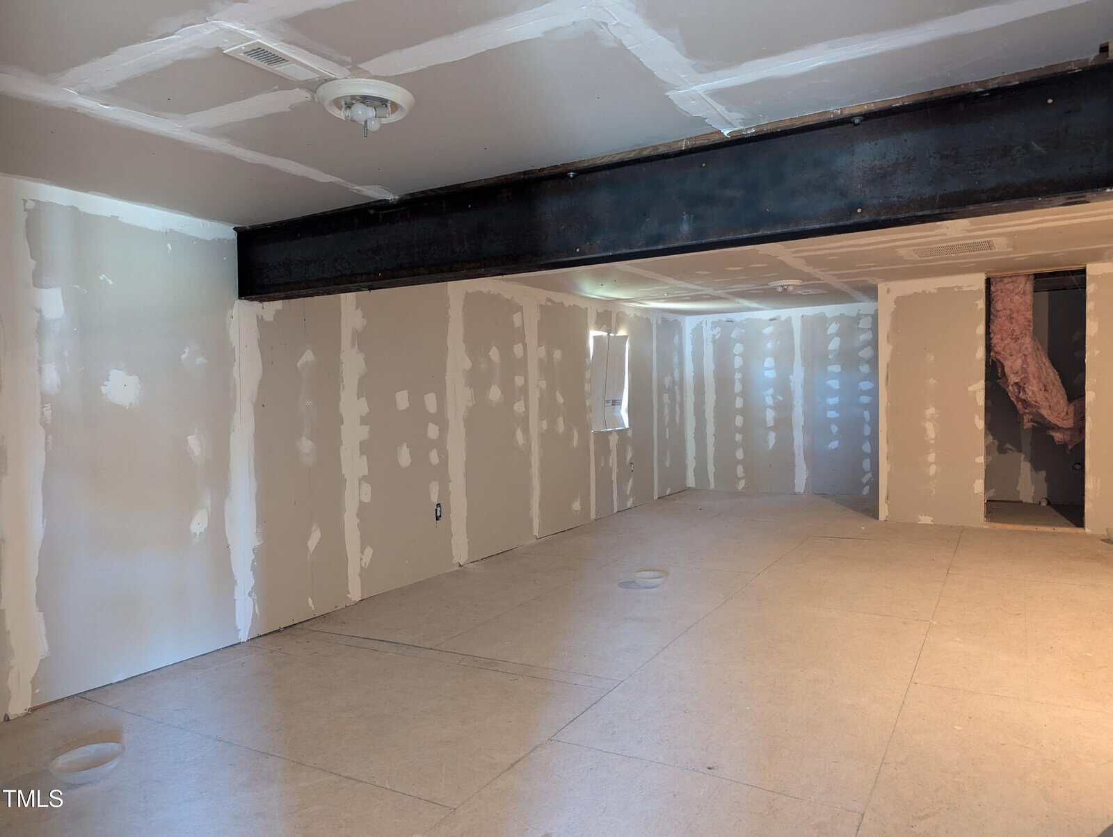 A spacious unfinished basement features drywall with visible seams and an overhead beam running across the ceiling. A small window allows some natural light to enter the room. An exposed area of insulation is visible at the far end.