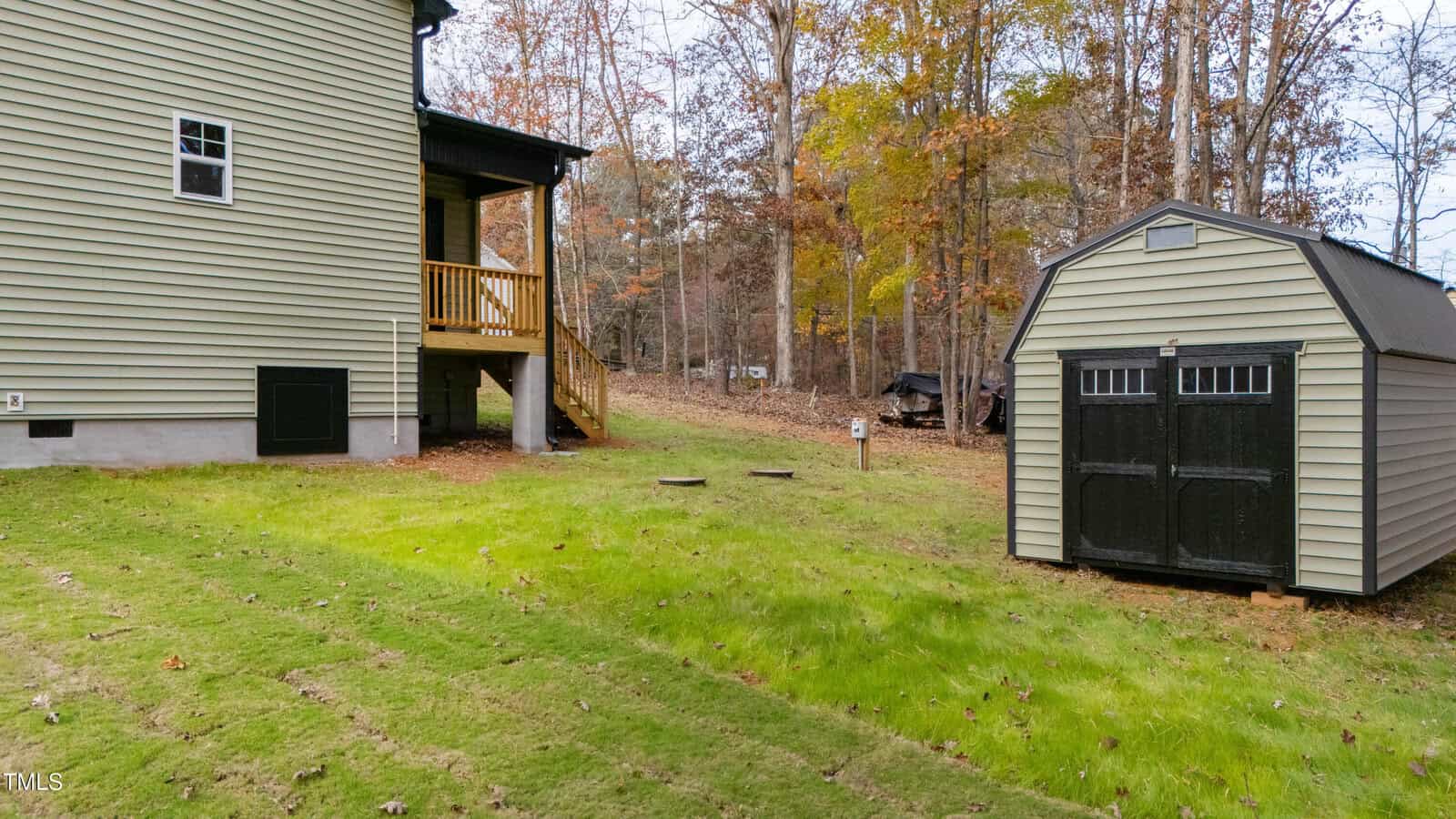 A light yellow house with a small wooden porch is on the left, and a beige shed with black double doors is on the right. The yard is grassy with some fallen leaves, bordered by woods with autumn foliage.