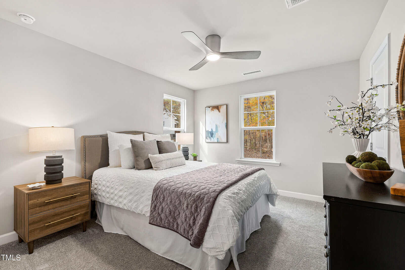 Bright bedroom with a neatly made bed, two windows, nightstand with a lamp, dresser with decor, ceiling fan, and neutral-colored walls and carpet. Artwork and a plant add decorative touches.
