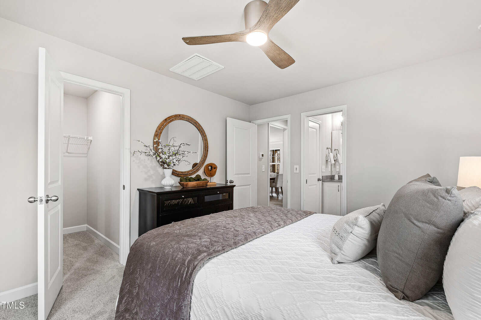 A modern bedroom with a ceiling fan, neatly made bed with gray and white bedding, a dark dresser with a round mirror, and an open door leading to a walk-in closet.