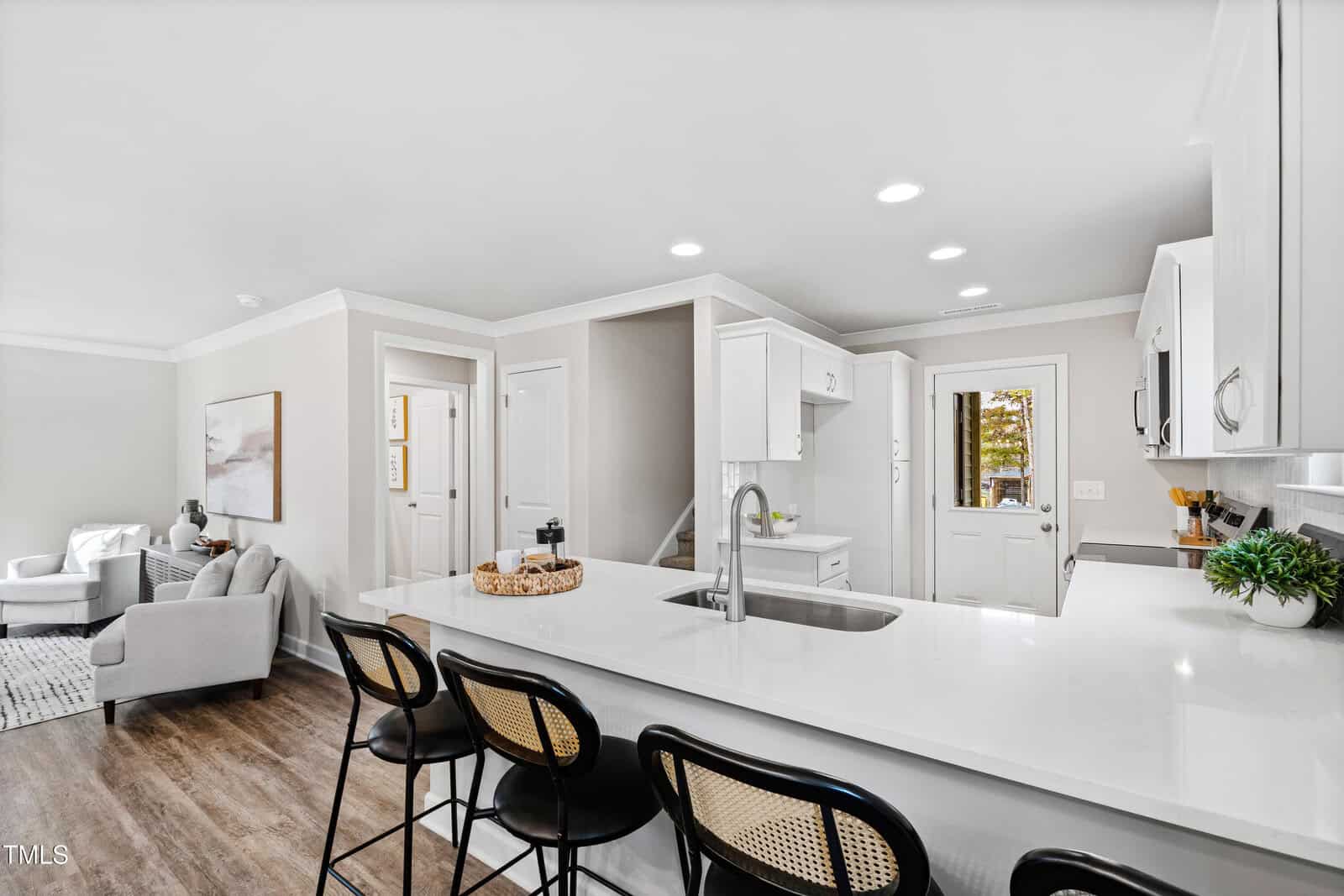 Modern kitchen with white cabinets and countertops, a sink in the island, three black barstools, and stainless steel appliances. The space opens to a living area with chairs and framed wall art. Wood flooring throughout.