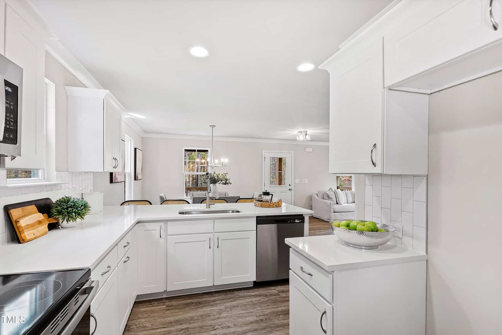 A modern kitchen with white cabinets, stainless steel appliances, white countertops, a subway tile backsplash, and wood-style flooring. A dining area and living room are visible in the background.