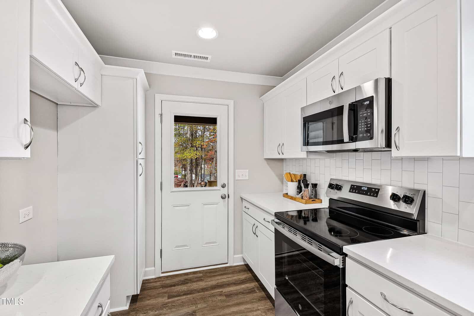 A small modern kitchen with white cabinets, stainless steel appliances, electric stove, microwave, white subway tile backsplash, wood-look flooring, and a door with a window leading outside.