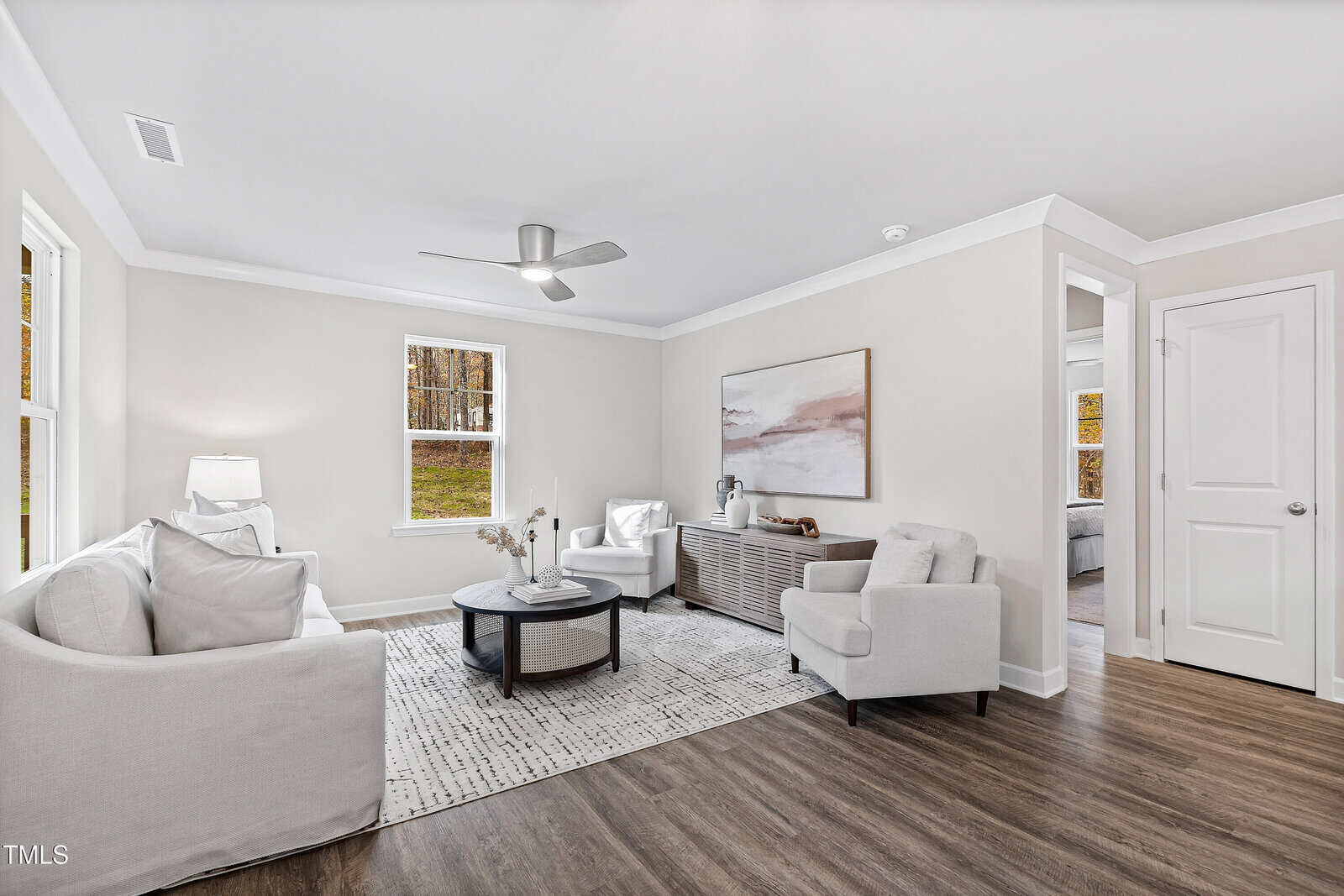A bright living room with white walls, wood flooring, and three white armchairs around a round coffee table. Large windows let in natural light. A sideboard and a large abstract painting are against the back wall.