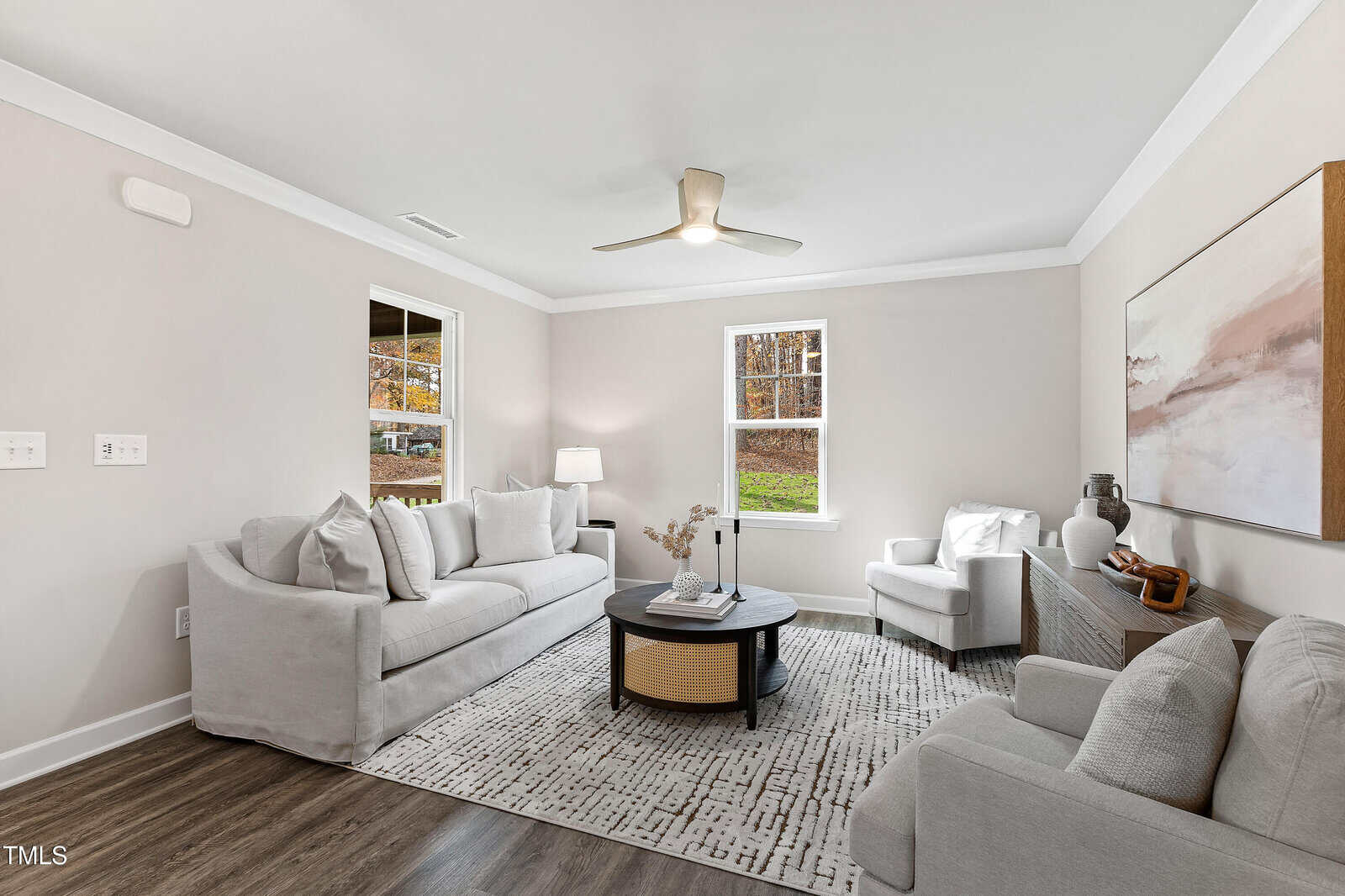 Modern living room with two light gray sofas, a white armchair, round coffee table, neutral rug, large abstract painting, and a ceiling fan. Sunlight streams in through two windows, illuminating the cozy space.