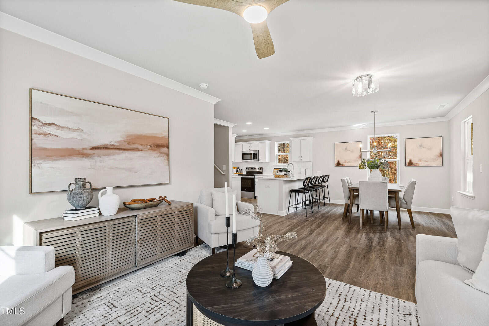 A modern open-concept living and dining area with neutral-toned furniture, a round coffee table, a large abstract painting, and a view into a white kitchen with a breakfast bar and three barstools.