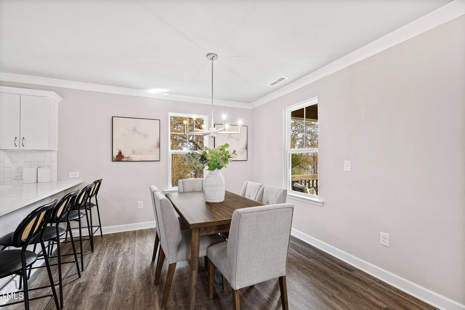 A modern dining area with a wooden table, six upholstered chairs, a vase with greenery, white walls, two windows, and part of a white kitchen with barstools on the left.