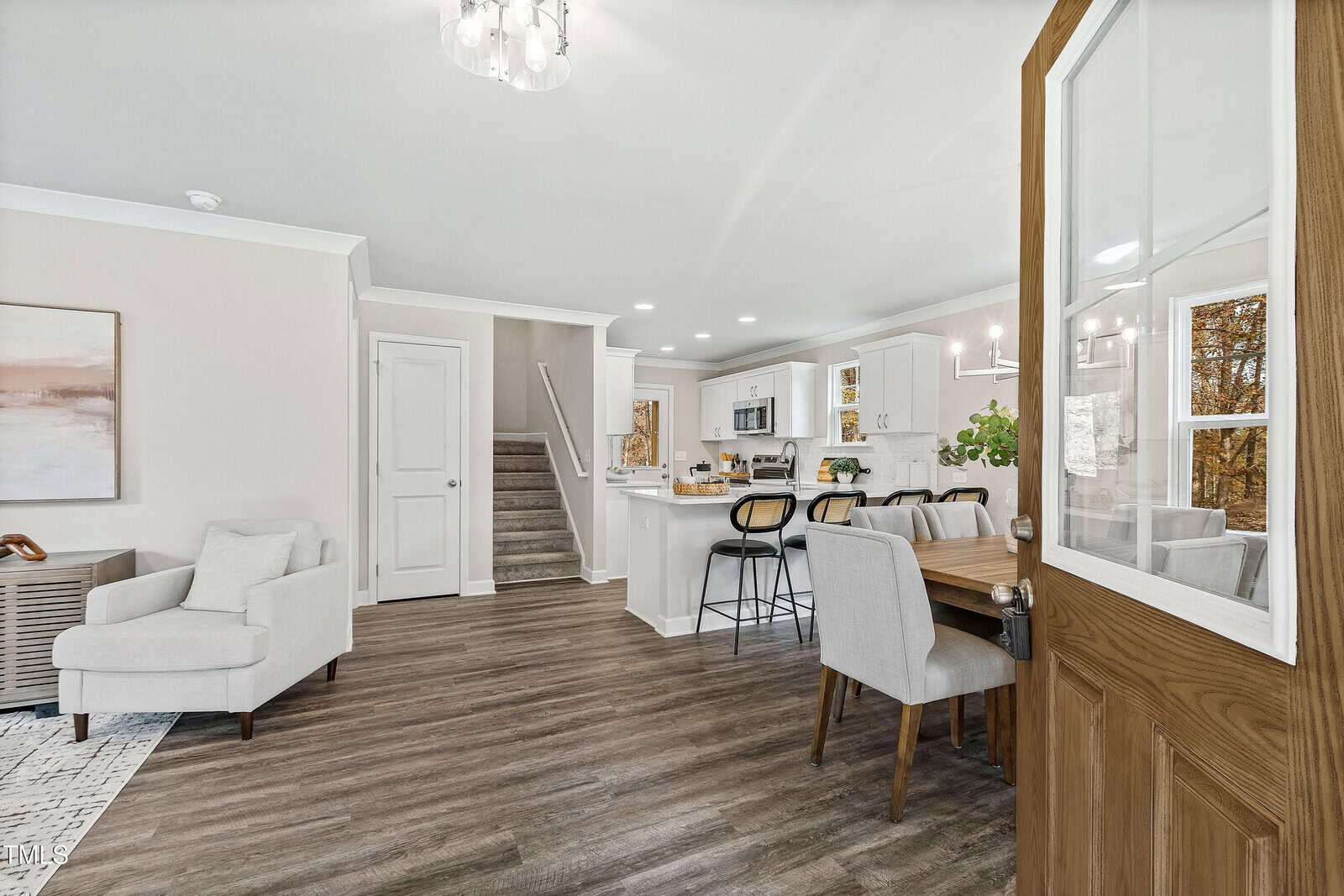 A modern open-concept living area with wood flooring, a white armchair, dining table with upholstered chairs, kitchen with white cabinets, a breakfast bar with stools, and a staircase leading upstairs.