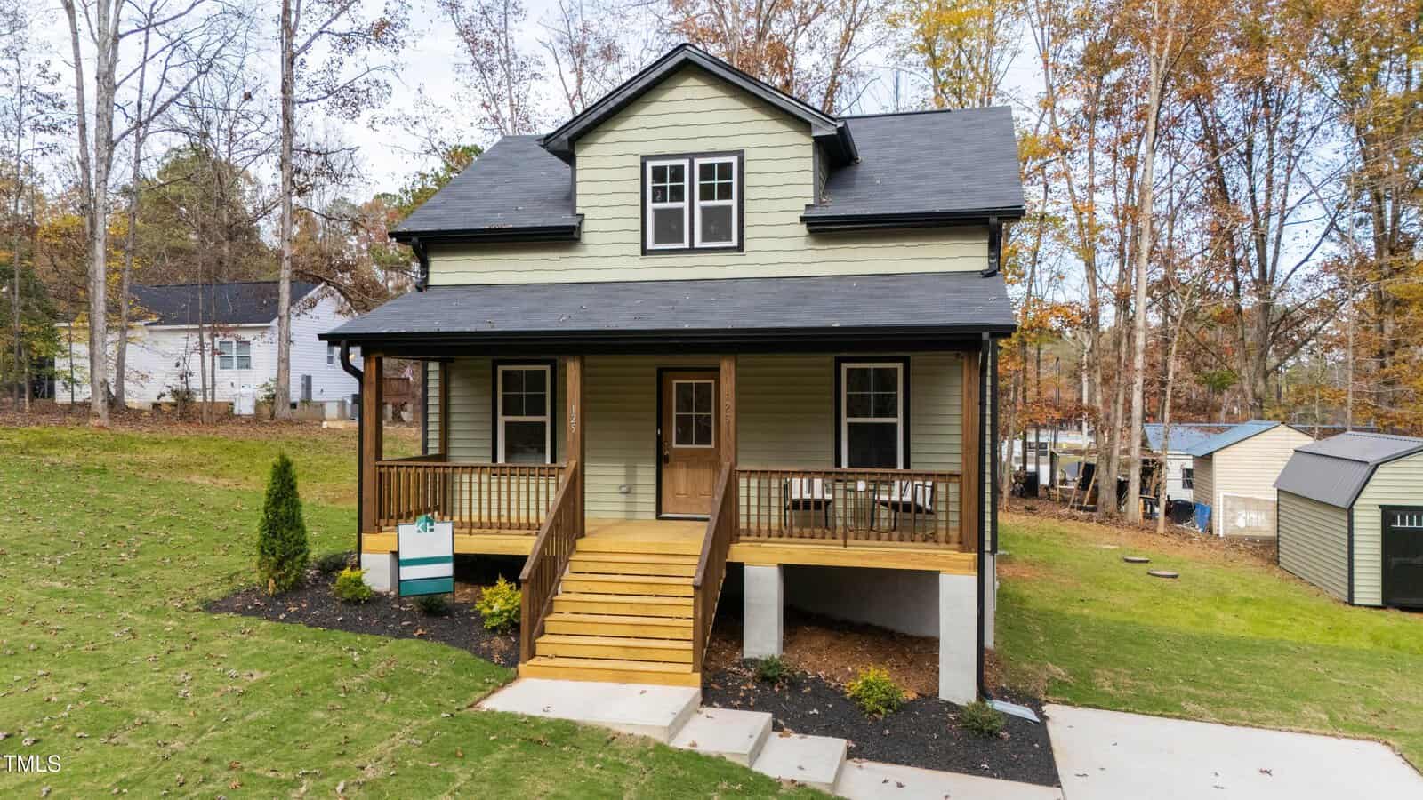 A two-story green house with a covered front porch, wooden railings, and steps leading up to the entrance. The house is surrounded by grass, trees, and neighboring houses, with sheds visible in the background.