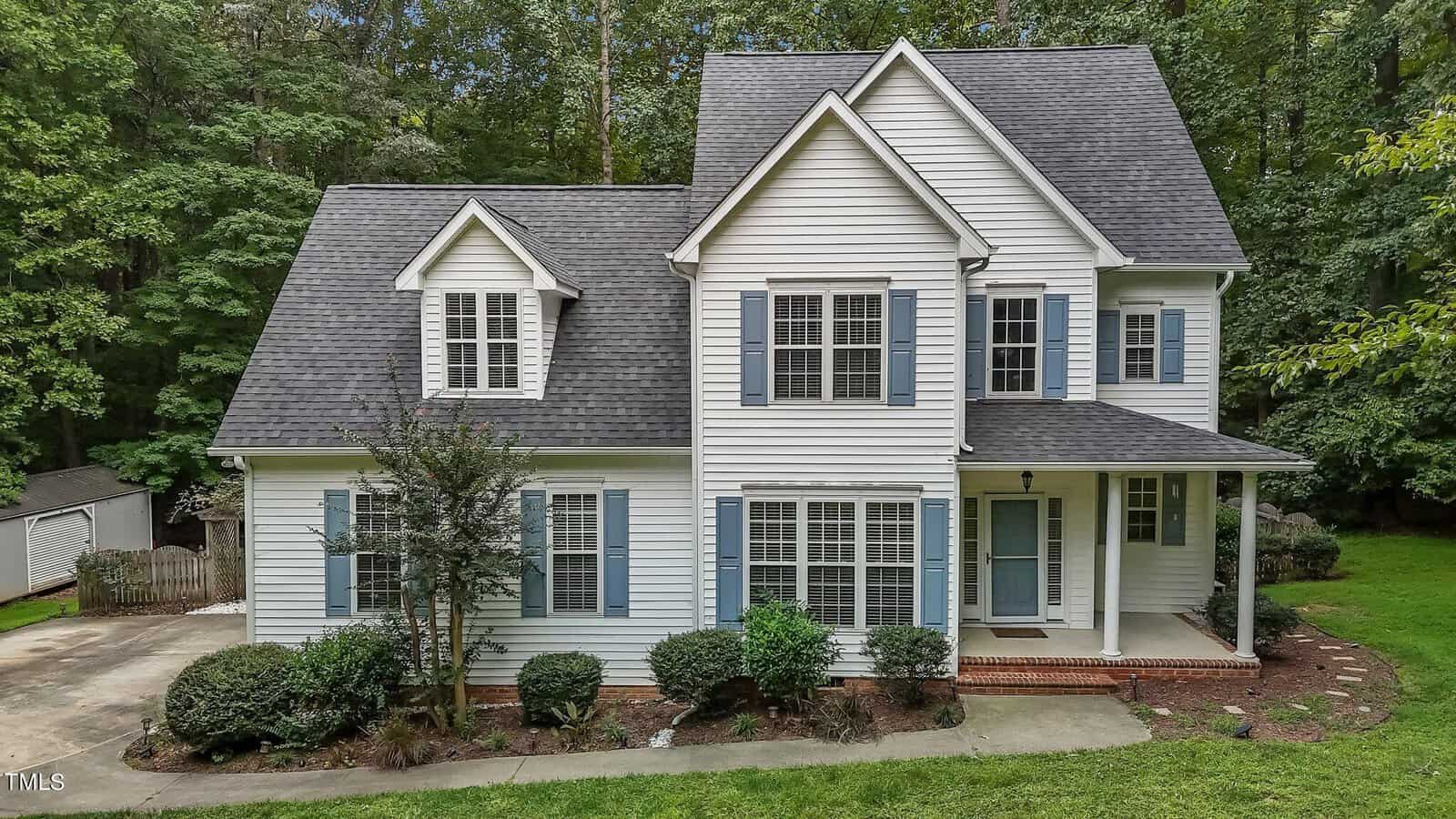 A two-story white house with blue shutters, a covered front porch, and a dark gray roof, surrounded by trees and greenery. There is a driveway and neatly trimmed bushes in the front yard.