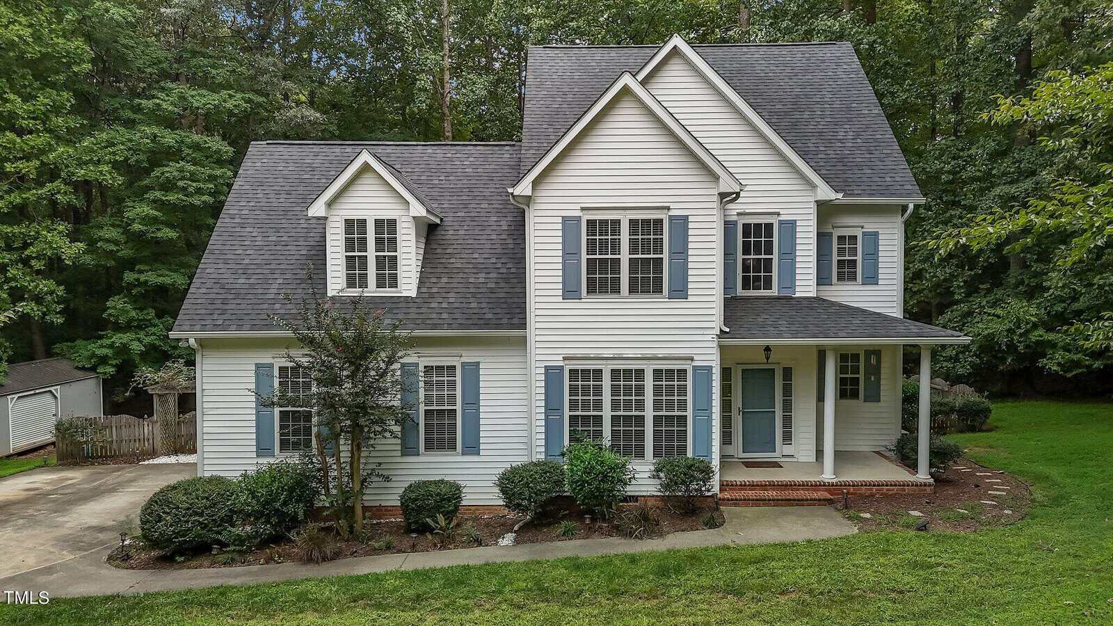 Two-story white house with blue shutters, gable roof, and a covered front porch. The house is surrounded by green grass, bushes, and tall trees in the background. There is a driveway to the left.