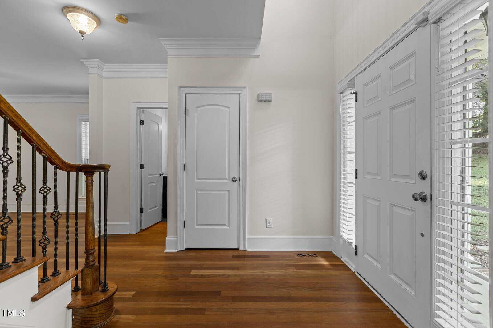 Bright entryway with wooden flooring, white walls, double front doors with windows, a staircase with a wooden handrail and iron balusters, and a closed interior door leading to another room.