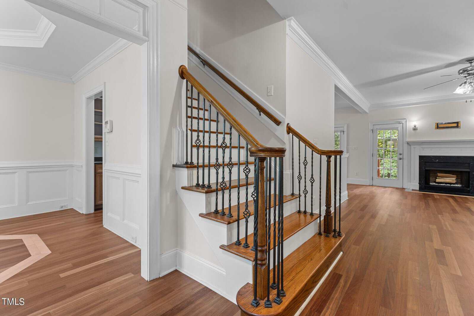 A wooden staircase with black iron balusters curves upward in a bright, spacious home interior with hardwood floors, white walls, crown molding, and a fireplace visible in the background.