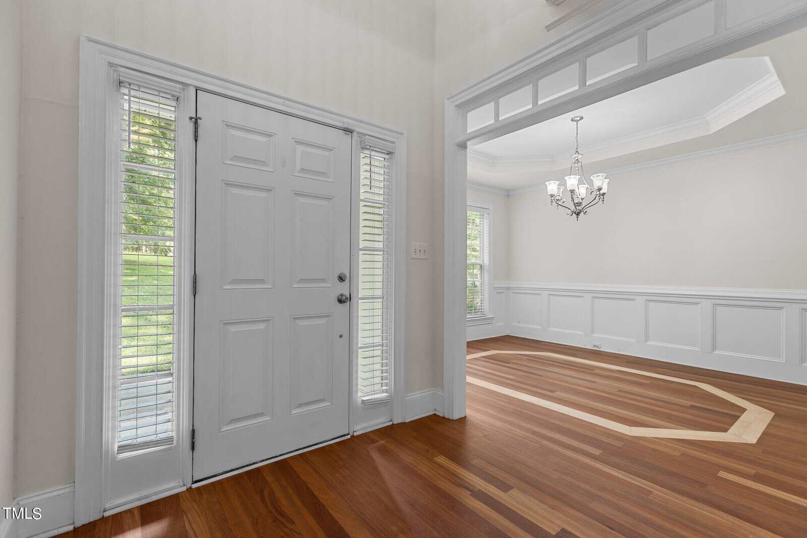 A bright entryway with white double doors, tall sidelights, and hardwood floors leads into an adjacent dining room with white wainscoting, large windows, and a decorative chandelier.