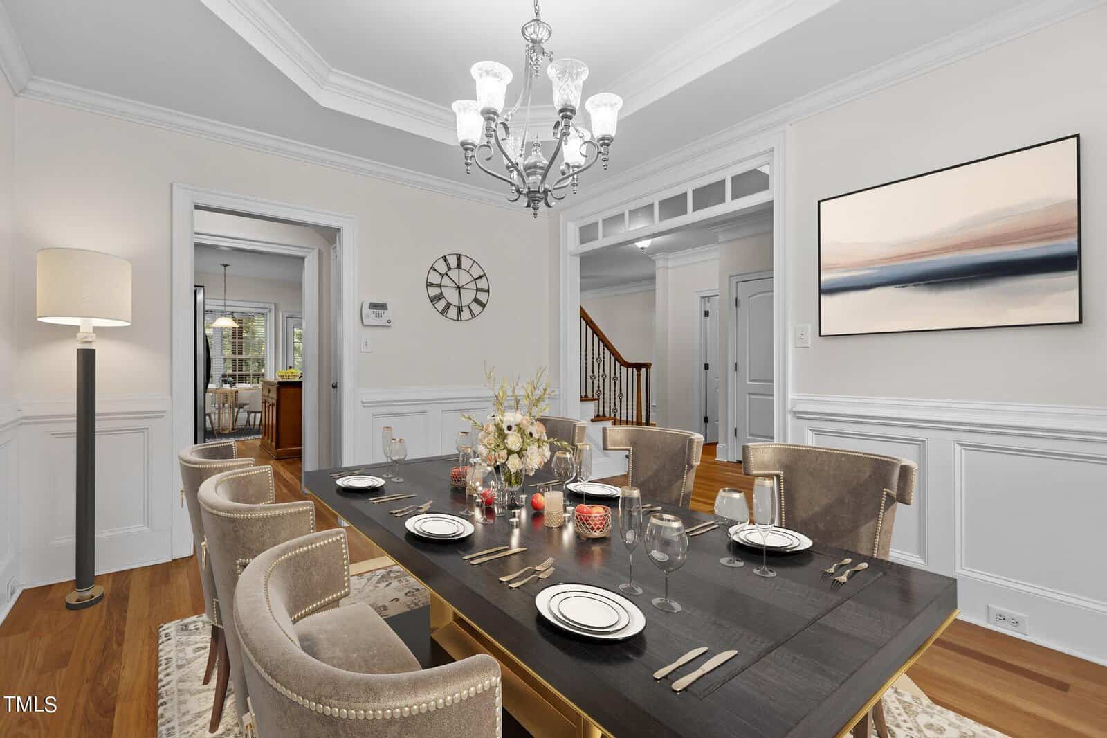 Elegant dining room with a dark wood table set for six, beige upholstered chairs, a floral centerpiece, and candles. A modern chandelier hangs above, and neutral art decorates the light-colored walls. A staircase is visible in the background.