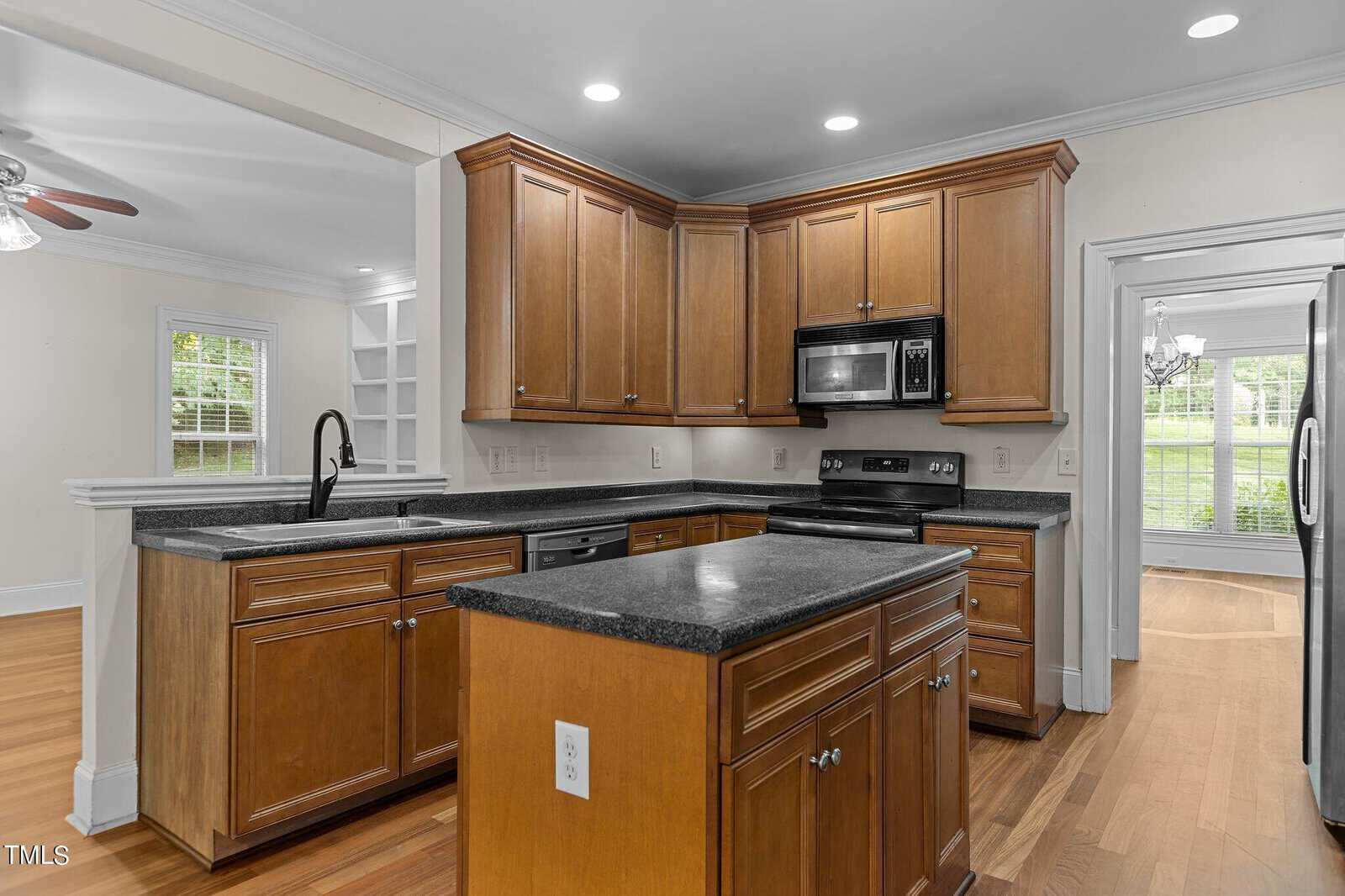 A modern kitchen with wooden cabinets, black countertops, an island, stainless steel appliances including a stove, microwave, and refrigerator, and an open layout with hardwood floors.