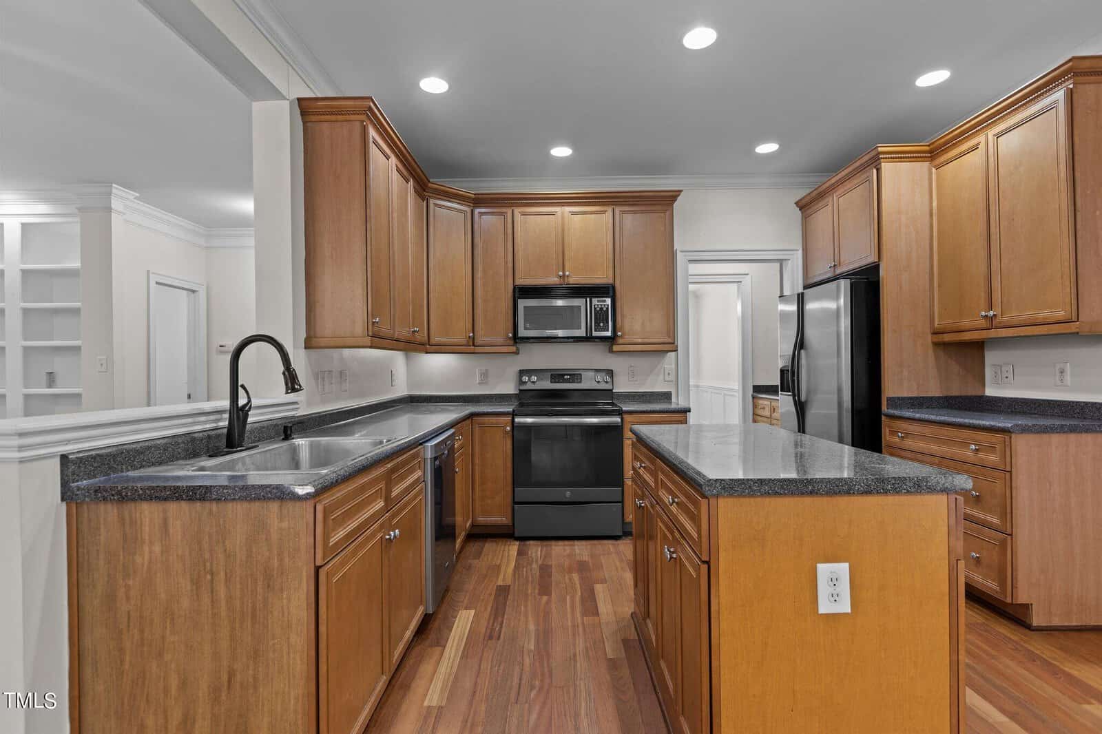Modern kitchen with wooden cabinets, black appliances, dark countertops, a central island with a power outlet, and a black faucet. Recessed ceiling lights and hardwood floors complete the space.