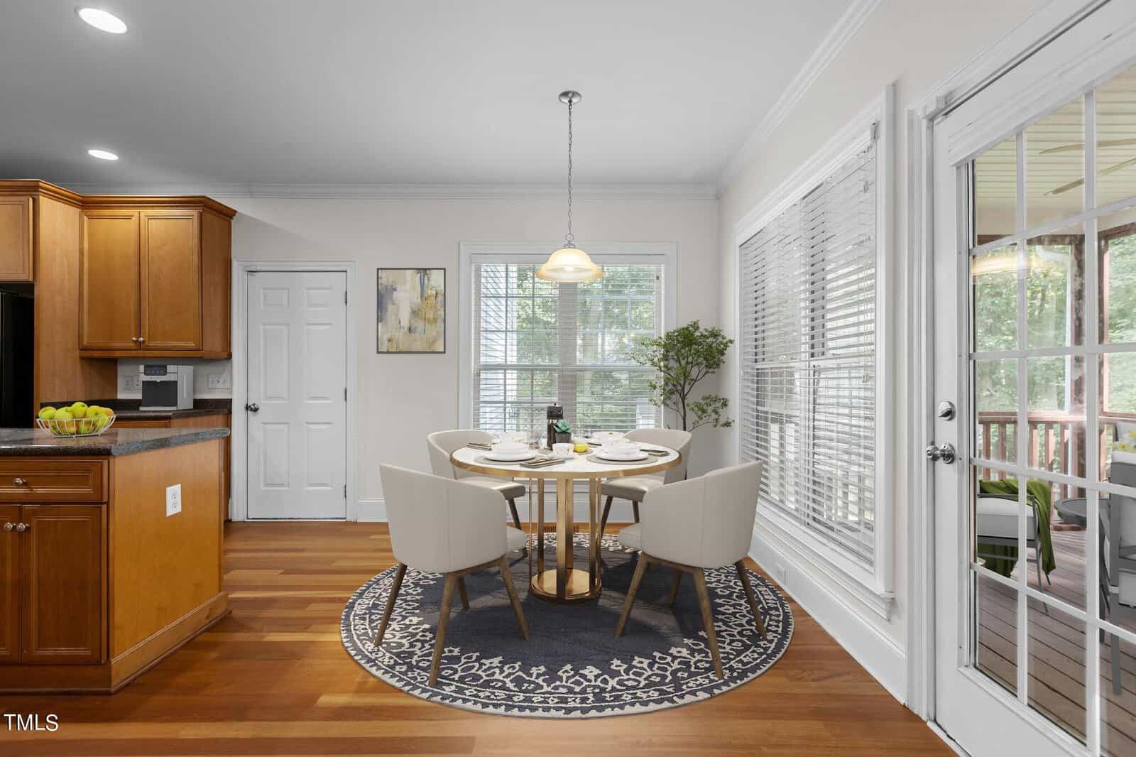 A modern dining area with a round table and four white chairs sits on a patterned rug by large windows. Wood floors, light walls, and a glass door lead to a porch with trees visible outside. Kitchen cabinets are on the left.
