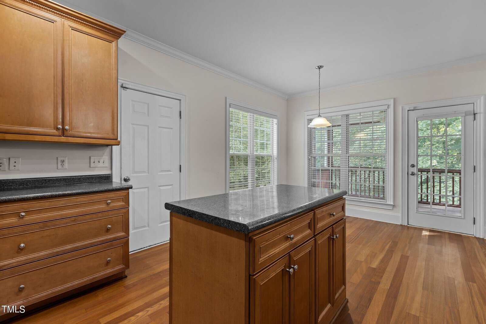 A bright kitchen with wood cabinets and floors, a central island with a dark countertop, white walls, large windows, and a glass door leading to a deck outside.