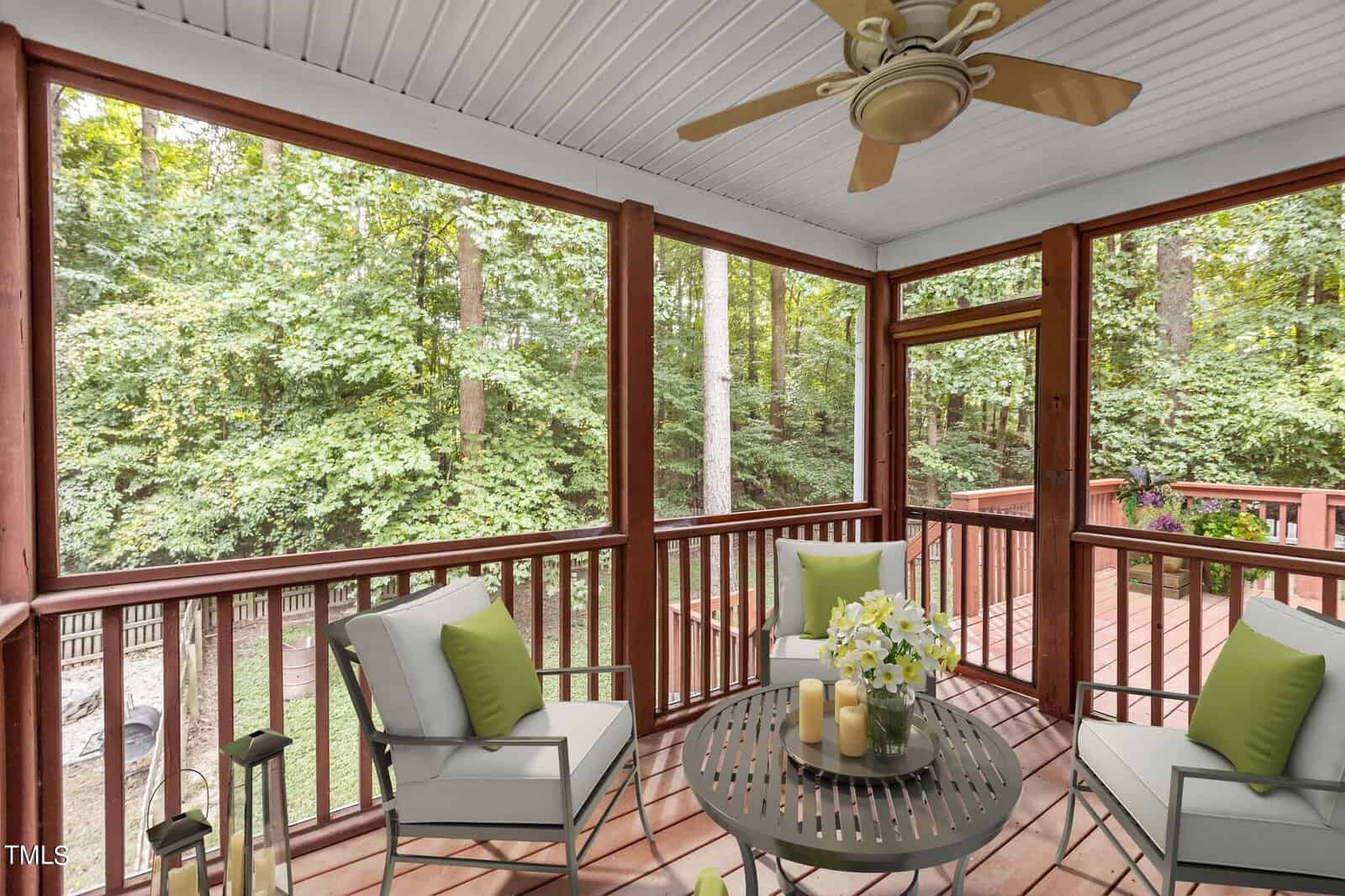 Screened-in porch with cushioned chairs, a round table with candles and flowers, ceiling fan, and wooded backyard view. Deck with railing visible through the screened panels.