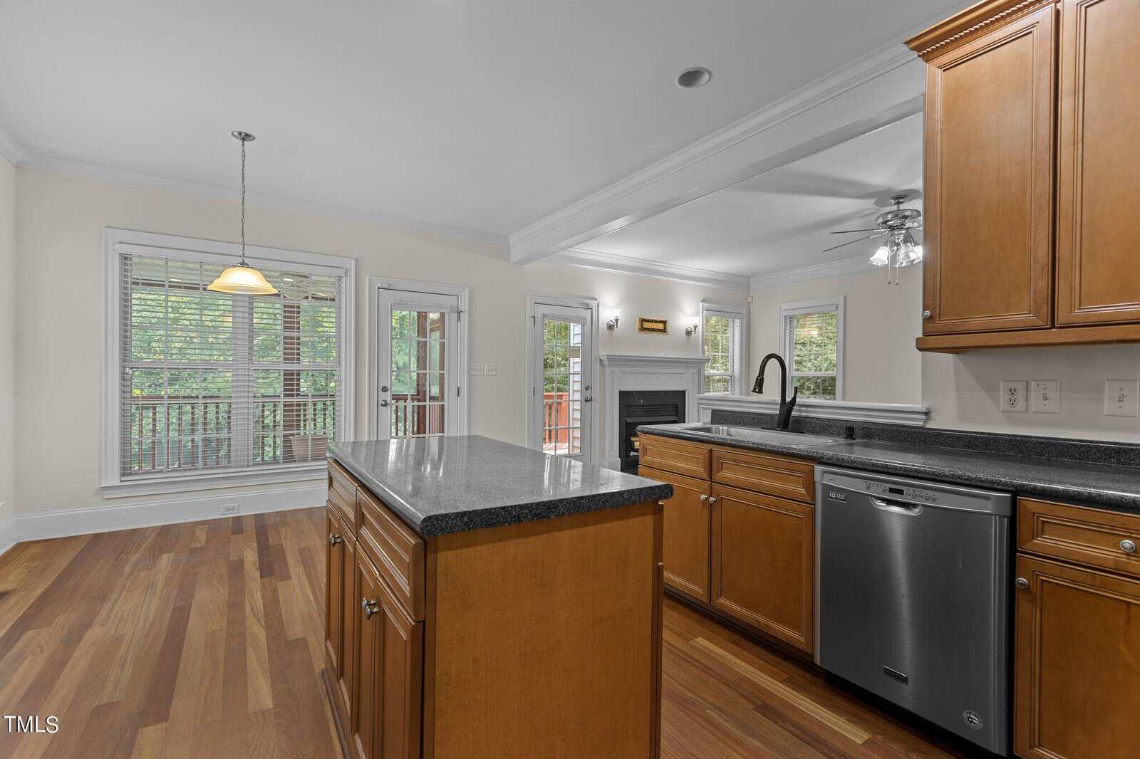 Modern kitchen with wood cabinets, black countertops, an island, stainless steel dishwasher, and sink. Open layout leads to a dining area, living room with fireplace, and large windows showing a deck and trees outside.