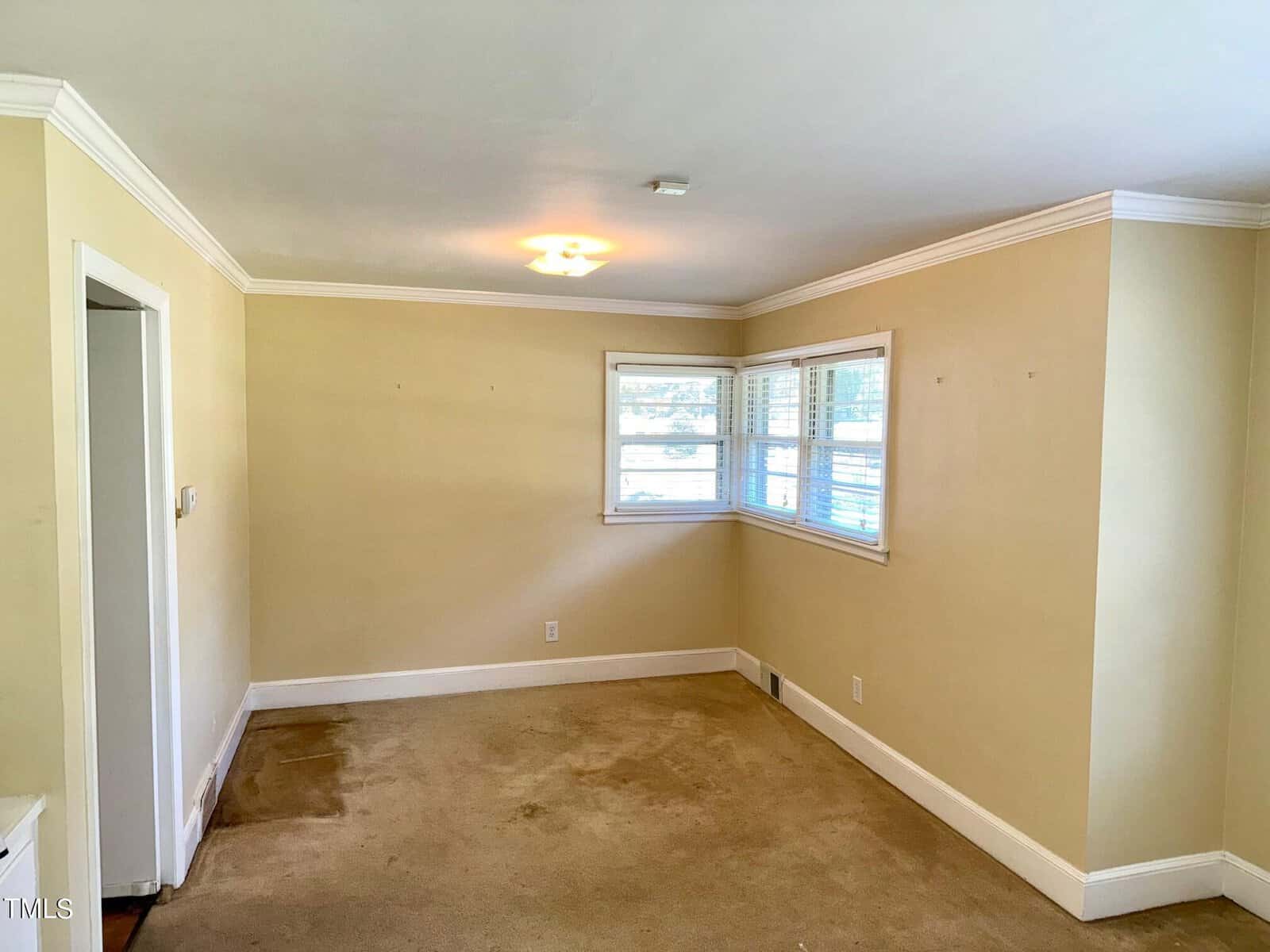 An empty room with beige walls and a pale carpet. The room features a central ceiling light and a window set in a corner, partially covered by white blinds. There's a doorway on the left side. The corners have crown molding, and there are some stains on the carpet.