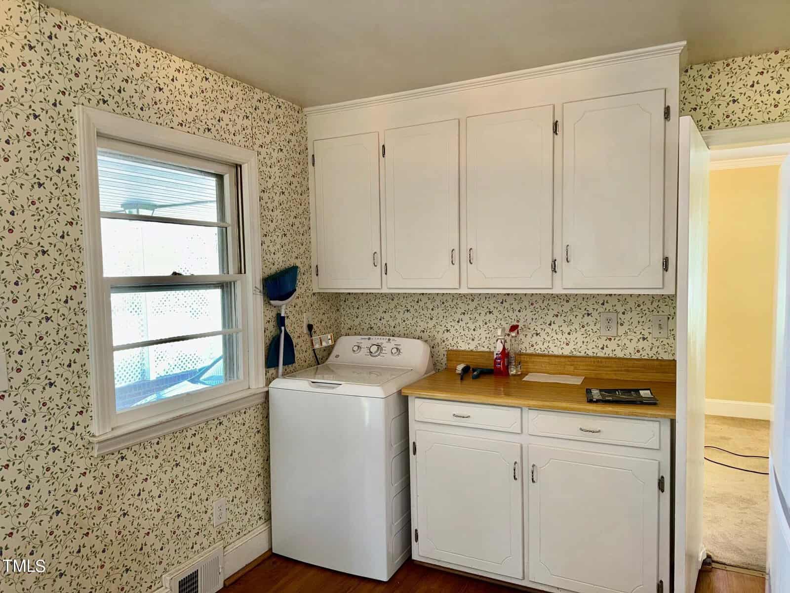 A small kitchen with floral wallpaper featuring white cabinetry, a washing machine, a window to the left, and a countertop holding a broom, cleaning supplies, and some tools. The room is well-lit with a view into an adjacent room with light-colored walls.