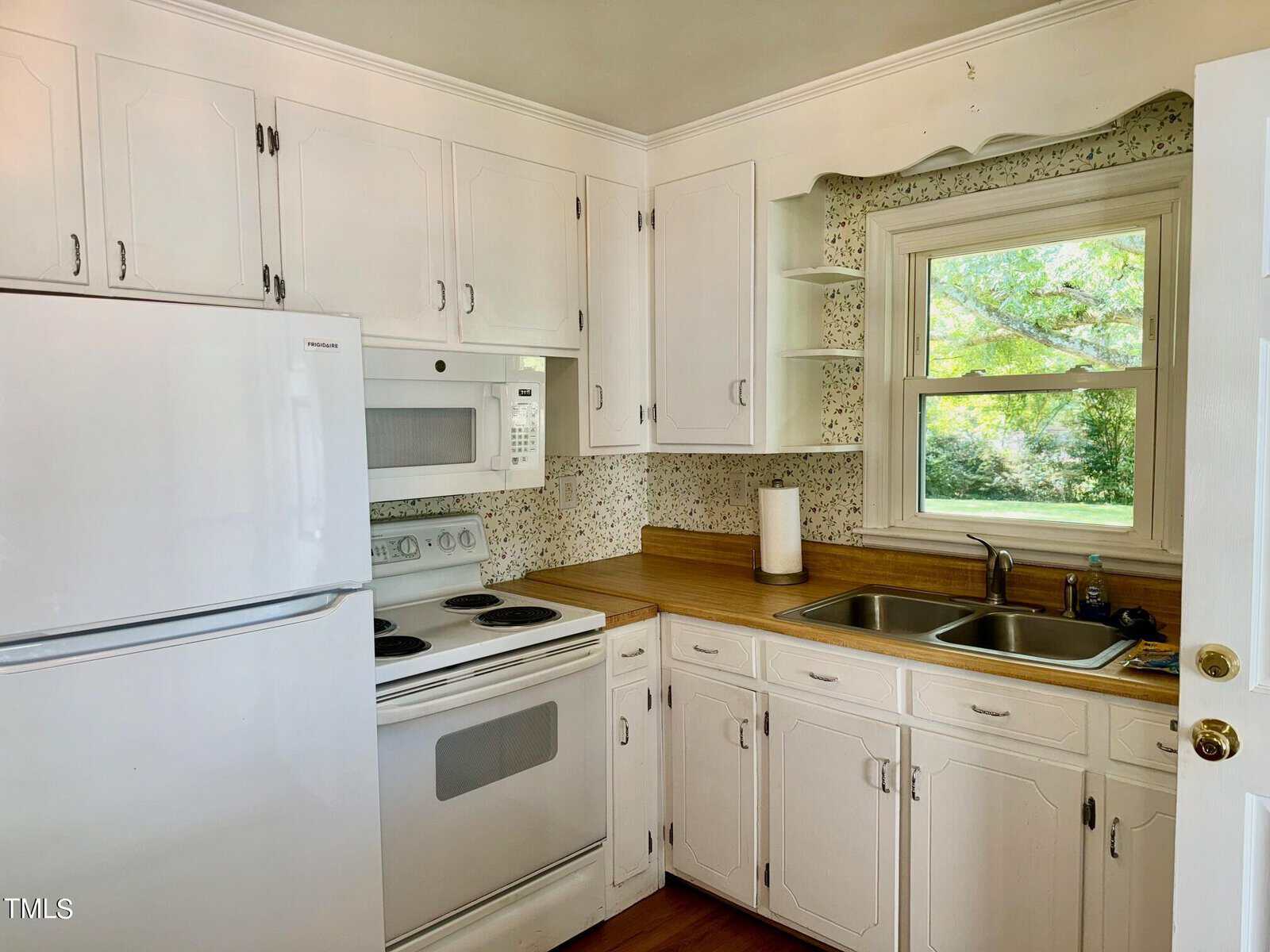 A small kitchen with white cabinets, a white refrigerator, oven, and microwave. There is a double sink by a window overlooking greenery. The countertops are wood-toned, and the backsplash features a subtle pattern. A paper towel holder is on the counter.
