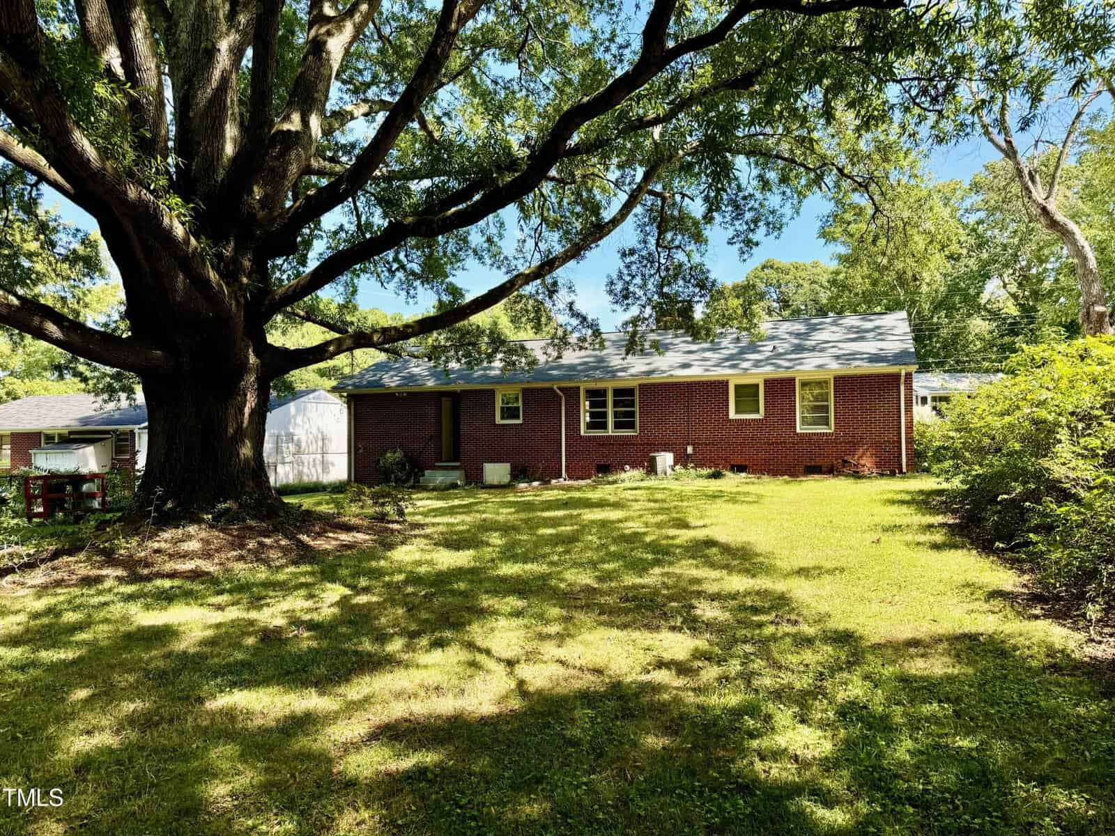 A brick single-story house with a gray roof, set within a lush backyard featuring a large tree providing shade. The yard is spacious and grassy, surrounded by various shrubs and trees. A white shed is partially visible on the left side near the house.