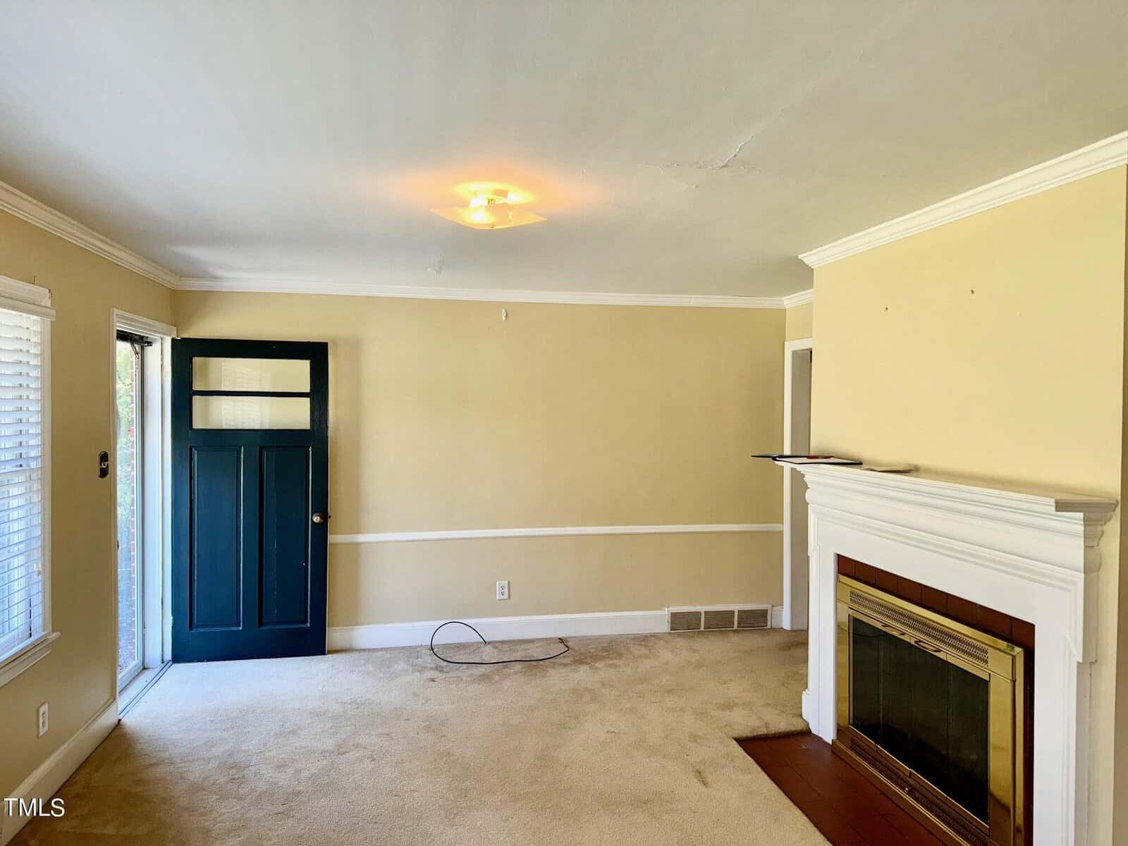 A beige-carpeted living room with light yellow walls. It features a white-framed fireplace with a dark interior on the right, a blue front door with glass panels on the left, and a ceiling light fixture. The room has a few white accents, including crown molding.