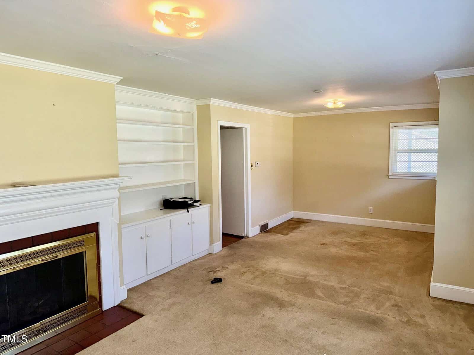 A living room with beige walls and carpet flooring. There is a white fireplace with a wooden mantel on the left, built-in white shelves and cabinets to the left of the fireplace, and a door in the middle leading to another room. Ceiling lights are on. A window is on the back wall.