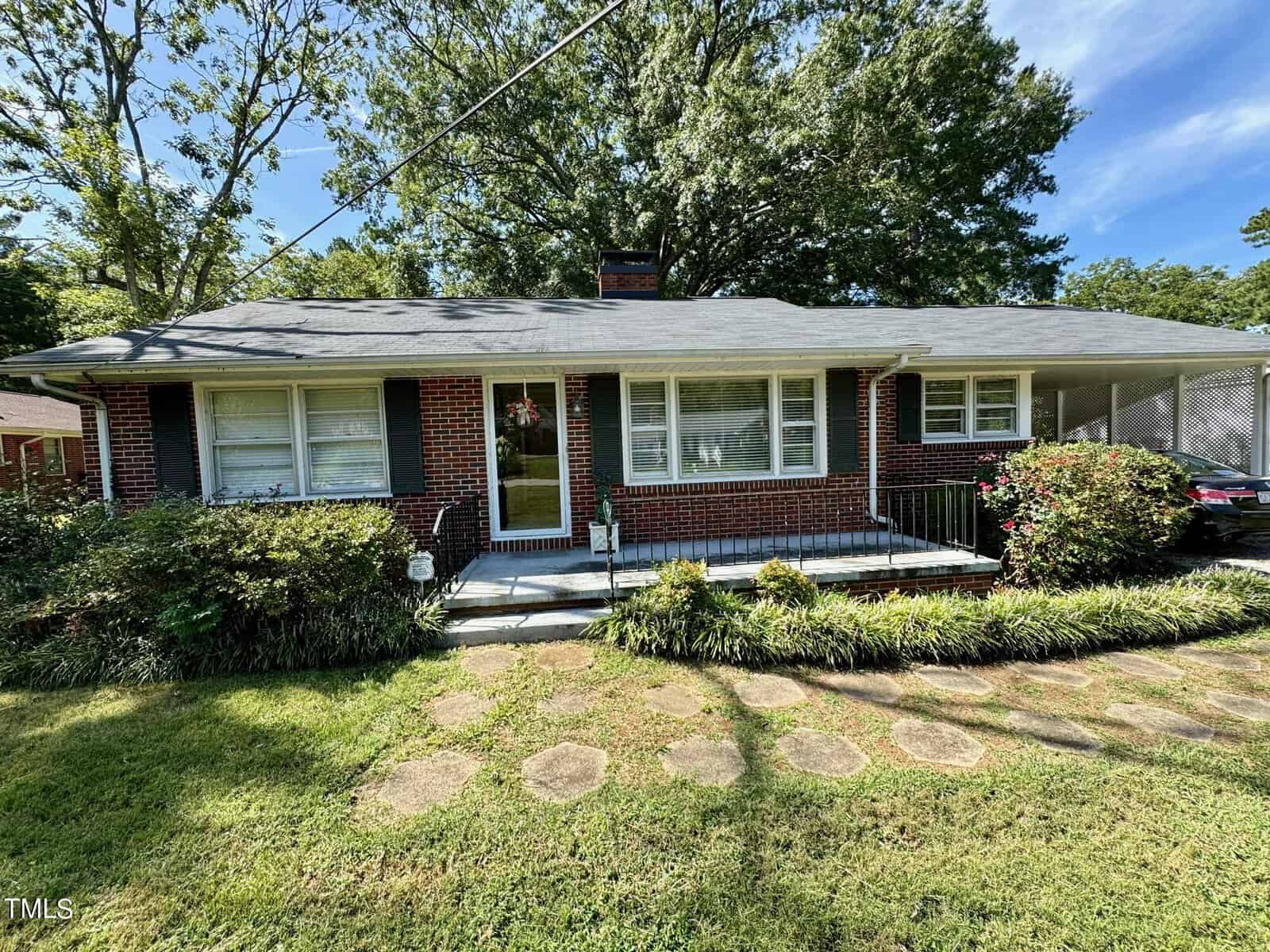 Located at 1205 Oakridge Avenue, this single-story brick house features a gray shingle roof, large front windows, and a central entrance. A concrete pathway leads to the front door, surrounded by landscaped bushes and grass. Large trees frame the yard, enhancing its charm under the clear Henderson sky.