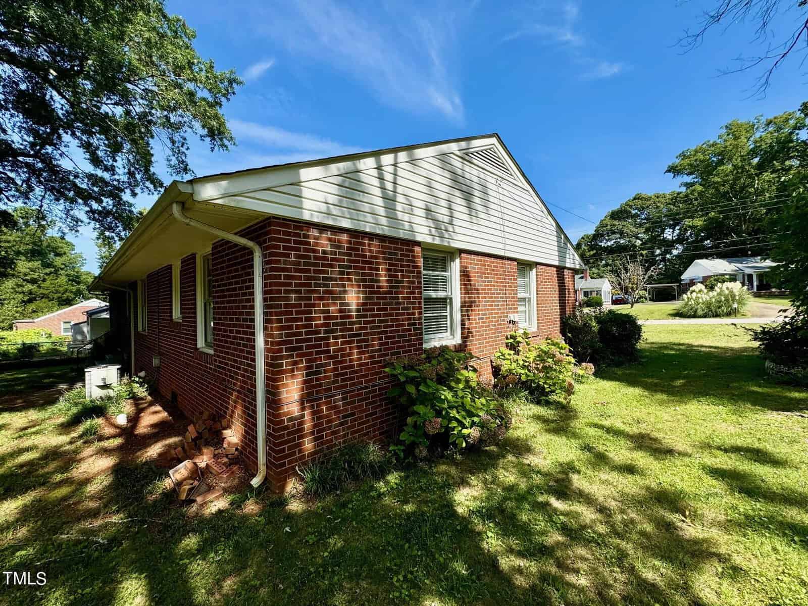 A brick single-story house with white siding on the upper portion, surrounded by a grassy yard with a few shrubs and trees. It's a sunny day with a clear blue sky. The house has several windows and is situated in a quiet neighborhood.