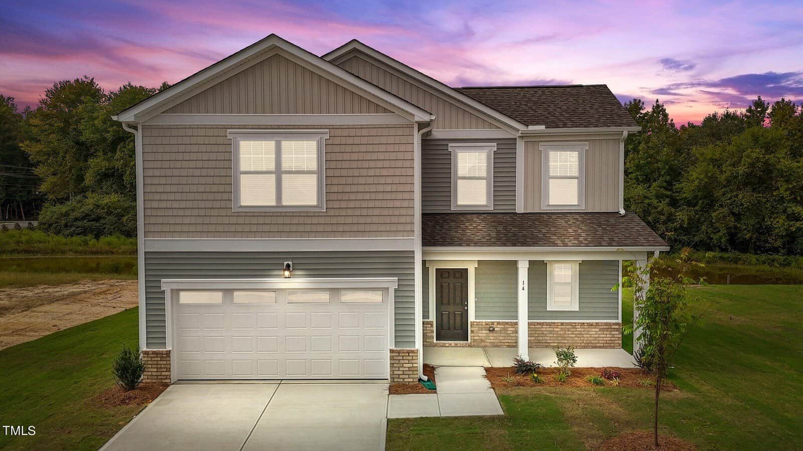 Two-story suburban house with gray siding, white trim, and a covered front porch. There is a double garage, a small front yard with young plants, and a driveway. Trees and sunset sky are in the background.