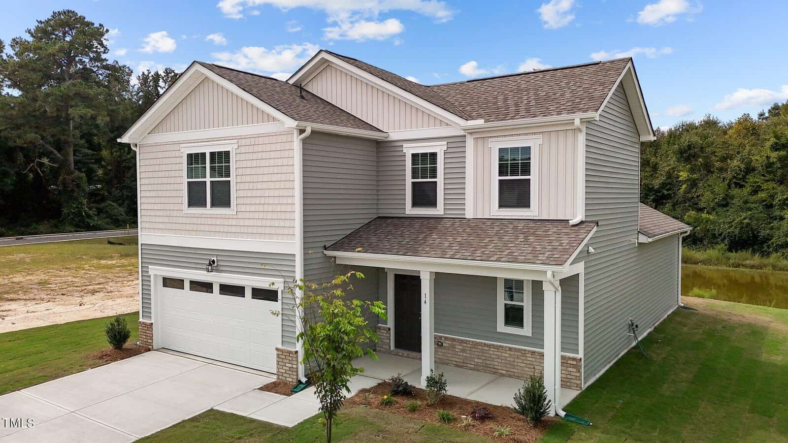 Two-story light gray house with attached two-car garage, white trim, brick accents, covered front porch, small shrubs in the yard, and a driveway, with trees and a pond in the background.