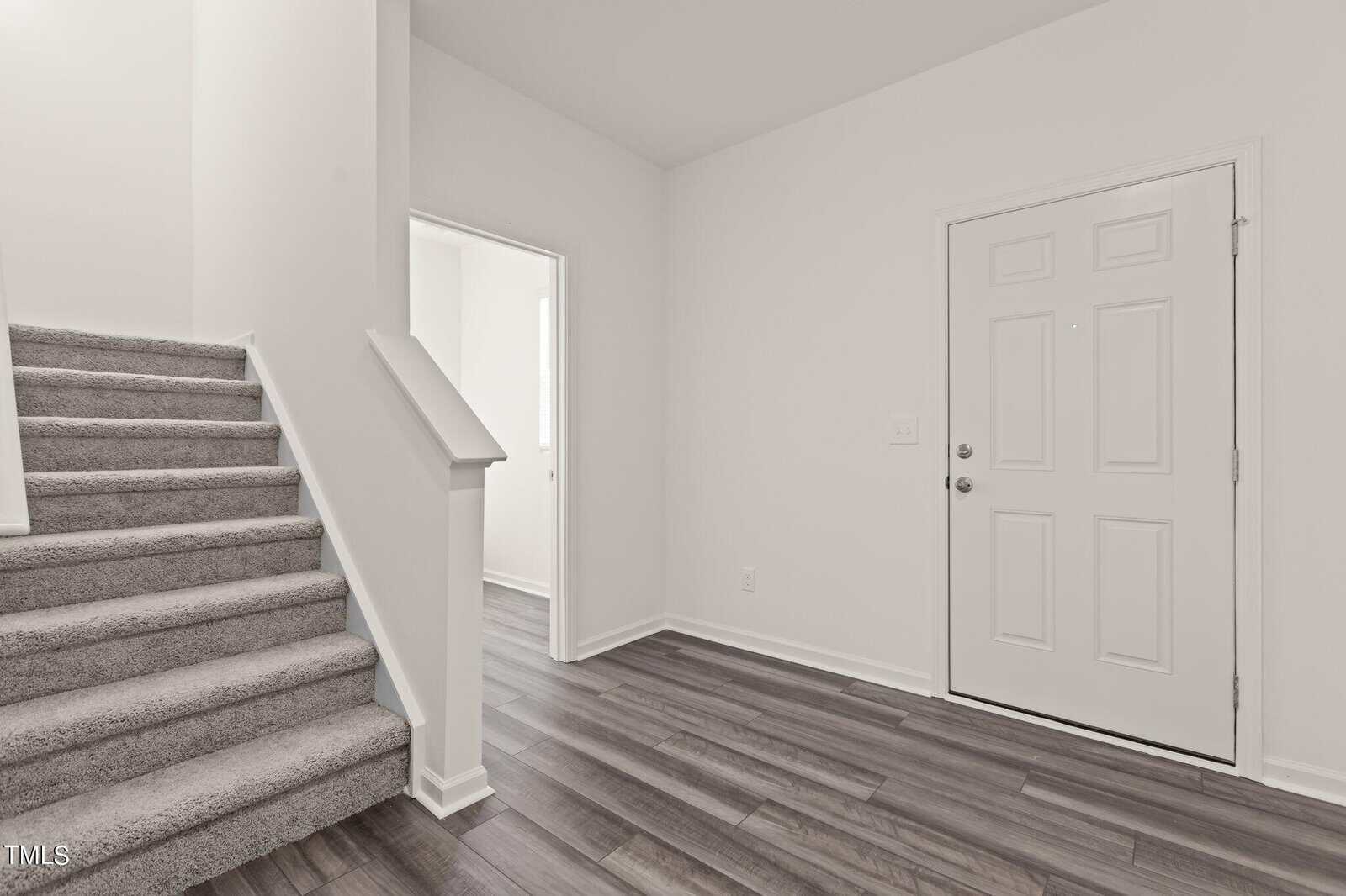 A white front door is on the right, with wood-look flooring and a carpeted staircase on the left. An open doorway leads to another well-lit room. The walls are plain white.