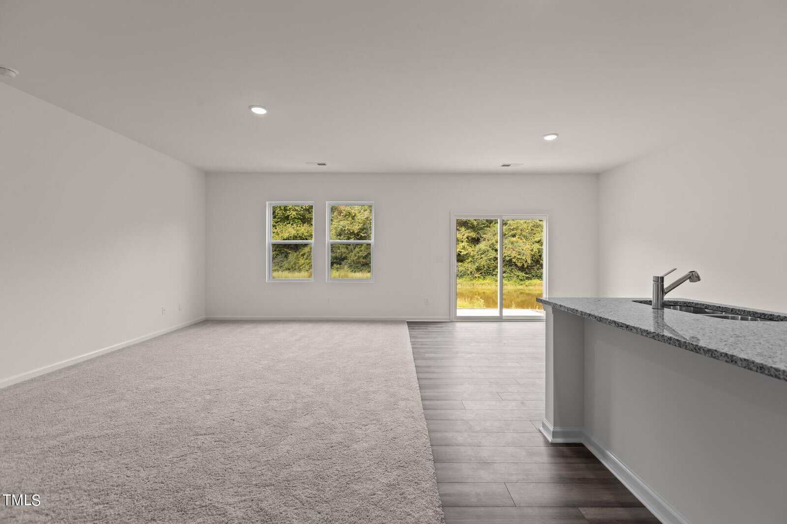 A spacious, empty room with light carpet and dark wood flooring, three windows, a sliding glass door leading outside, white walls, and a kitchen counter with a sink on the right.