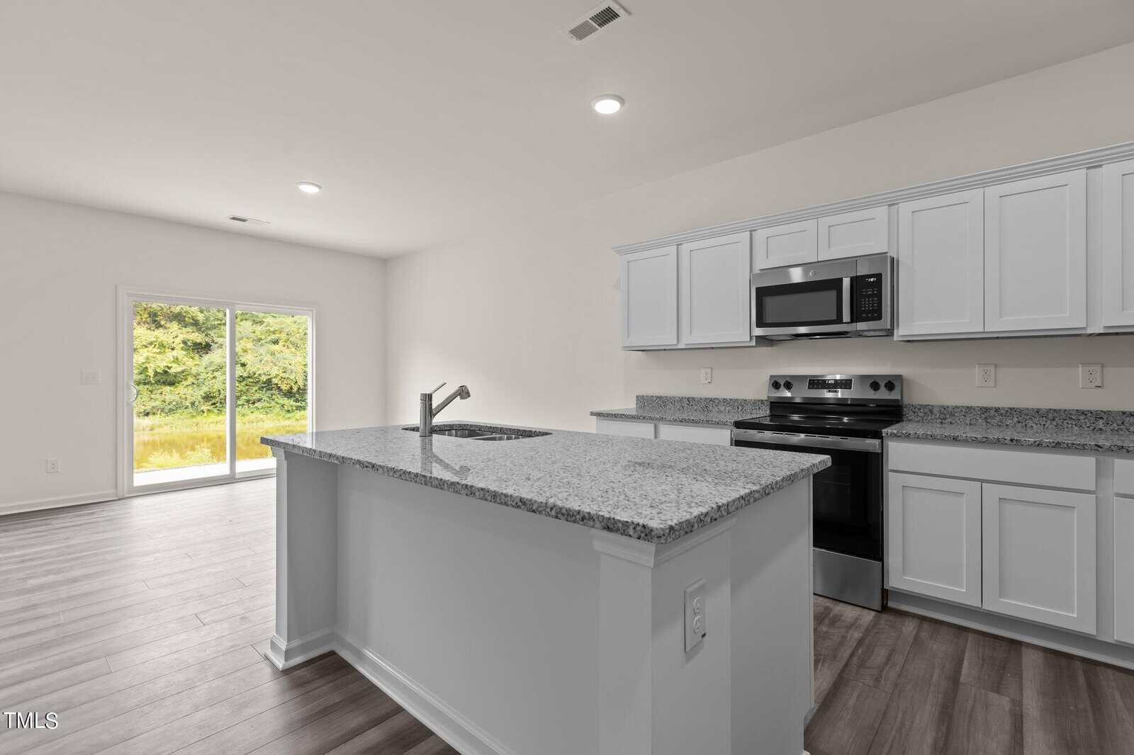 Modern kitchen with white cabinets, granite countertops, stainless steel appliances, and an island with a sink. Large glass sliding doors in the adjacent dining area provide a view of greenery outside.