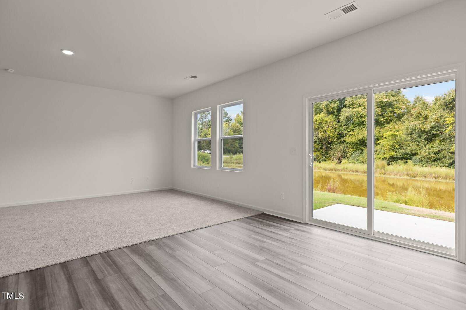 A bright, empty room with light wood flooring, carpeted section, two windows, and a sliding glass door leading to a patio with a view of grass, trees, and a pond. Walls and ceiling are white.