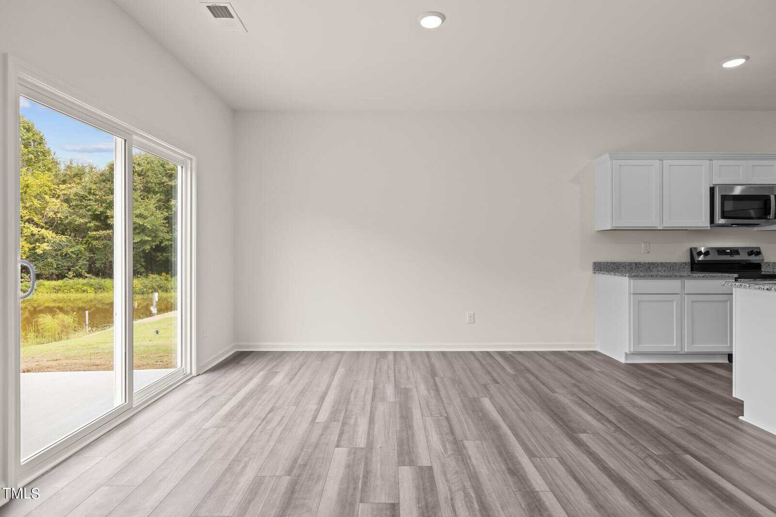 Bright, empty room with light gray wood floors, white walls, a large sliding glass door overlooking a grassy area, and a partial view of a kitchen with white cabinets and stainless steel appliances.