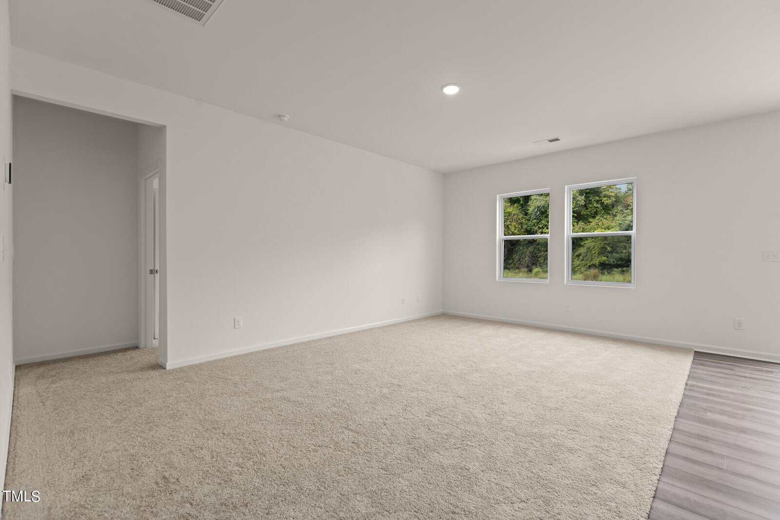 Spacious empty room with beige carpet, white walls, two windows showing greenery outside, and a section of wood flooring. A doorway leads to another area.