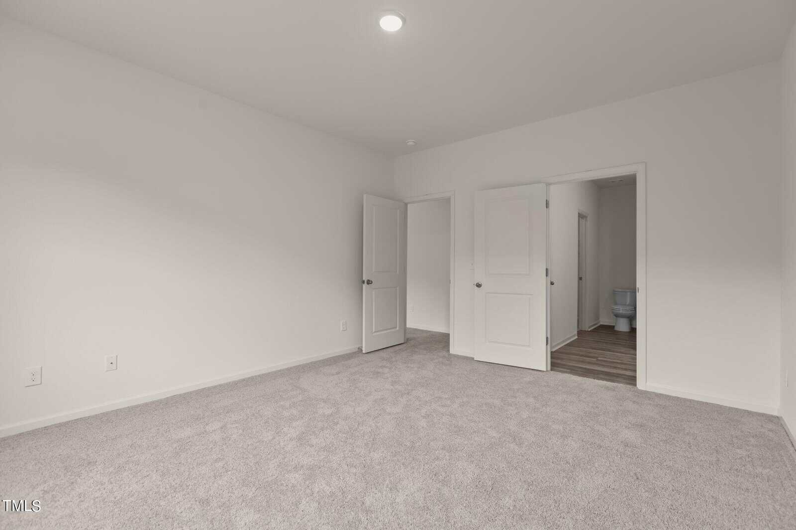 A bright, empty room with light gray carpet, white walls, and two open doors leading to another area with wood flooring and a partially visible toilet.