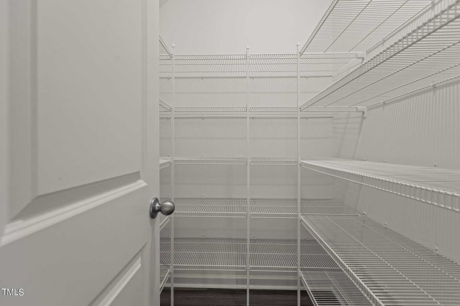 A walk-in pantry with white wire shelving units lining both sides and the back wall. The door is partially open, revealing empty shelves and a clean, neutral-colored interior.