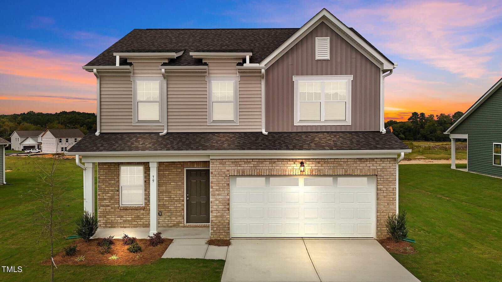 A two-story suburban house with gray siding and brick facade, white trim, double garage, and a neatly landscaped front yard, set against a colorful sunset sky.