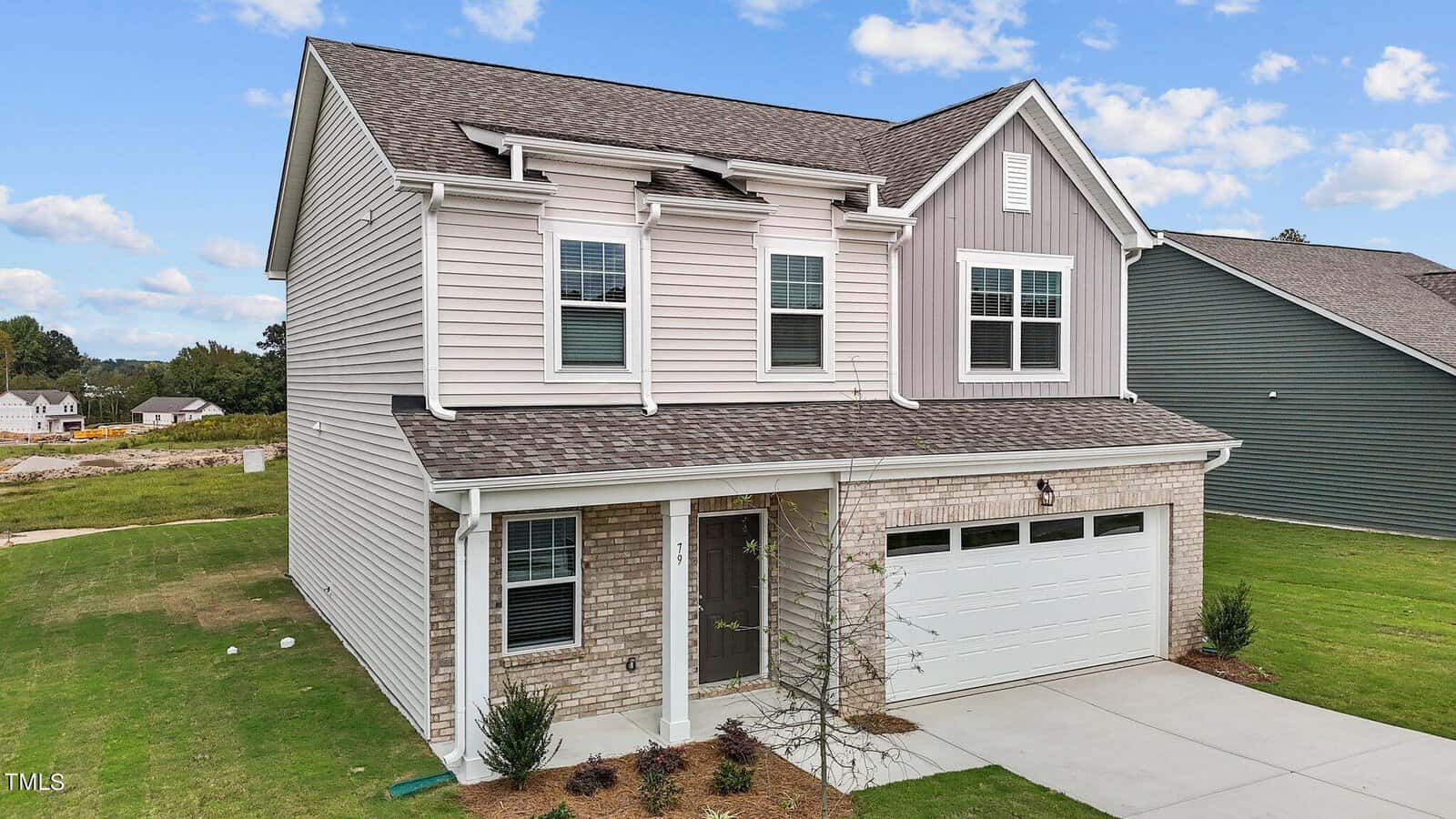 Two-story suburban house with beige siding, gray accents, and a brick facade. A double garage and small front porch are visible, surrounded by a green lawn and minimal landscaping under a partly cloudy sky.