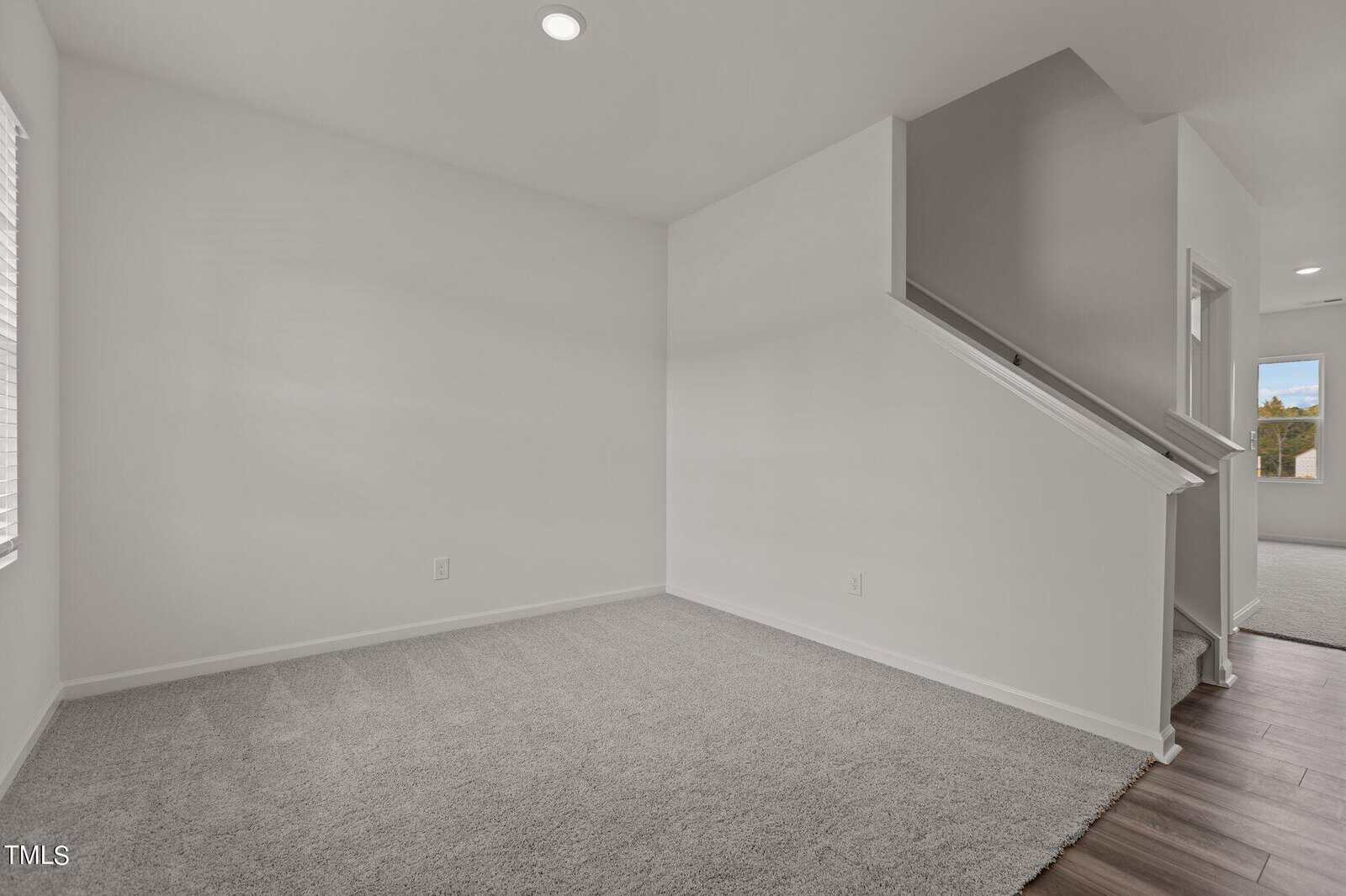 A bright, empty room with light gray carpet, white walls, a window on the left, and an open staircase leading to another area of the house with wood flooring.