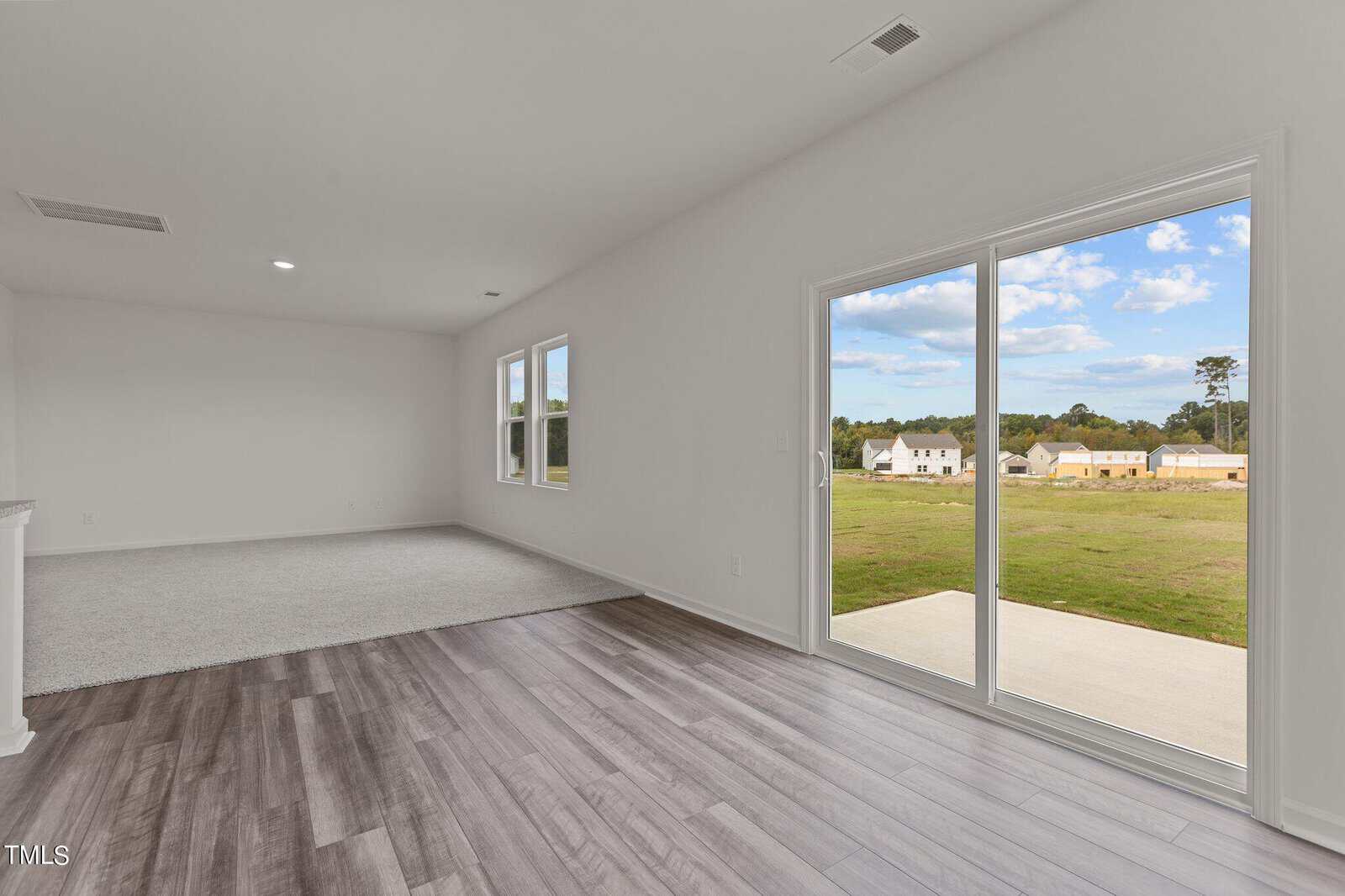 A modern empty room with light gray wood flooring, carpeted area, white walls, and large sliding glass doors opening to a patio and grassy backyard with houses in the distance under a partly cloudy sky.