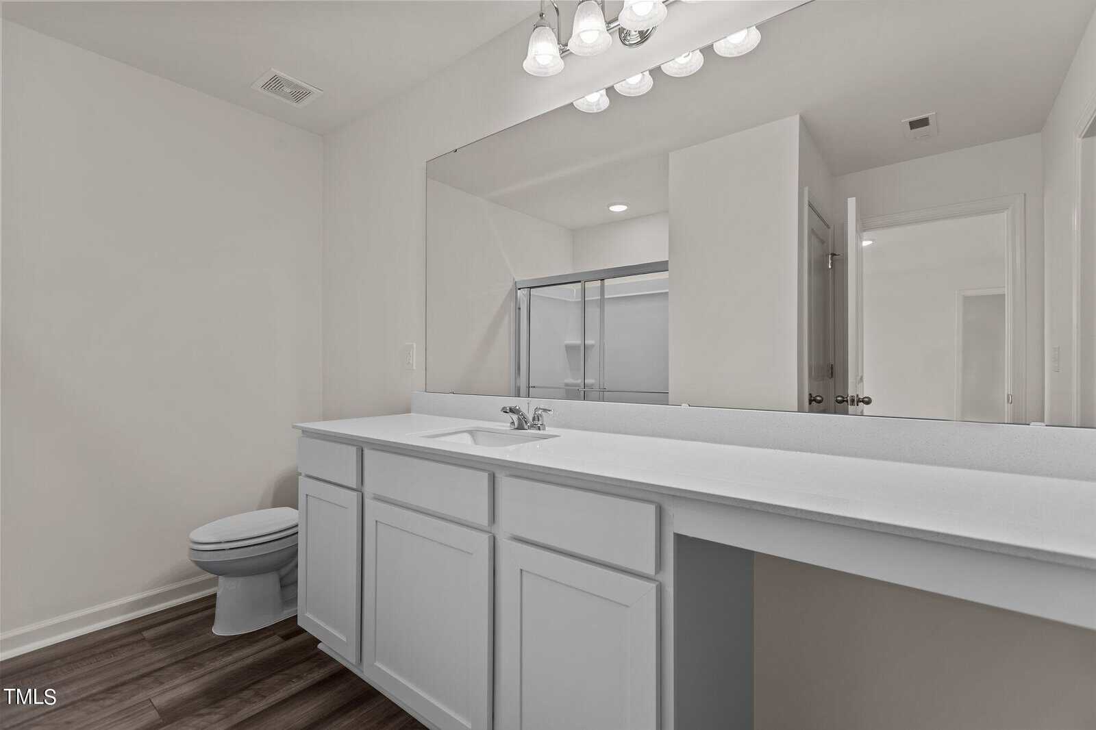 Modern bathroom with white cabinets, a wide countertop, single sink, toilet, large wall mirror, wood-style flooring, and a shower with glass doors in a bright, minimalistic design.