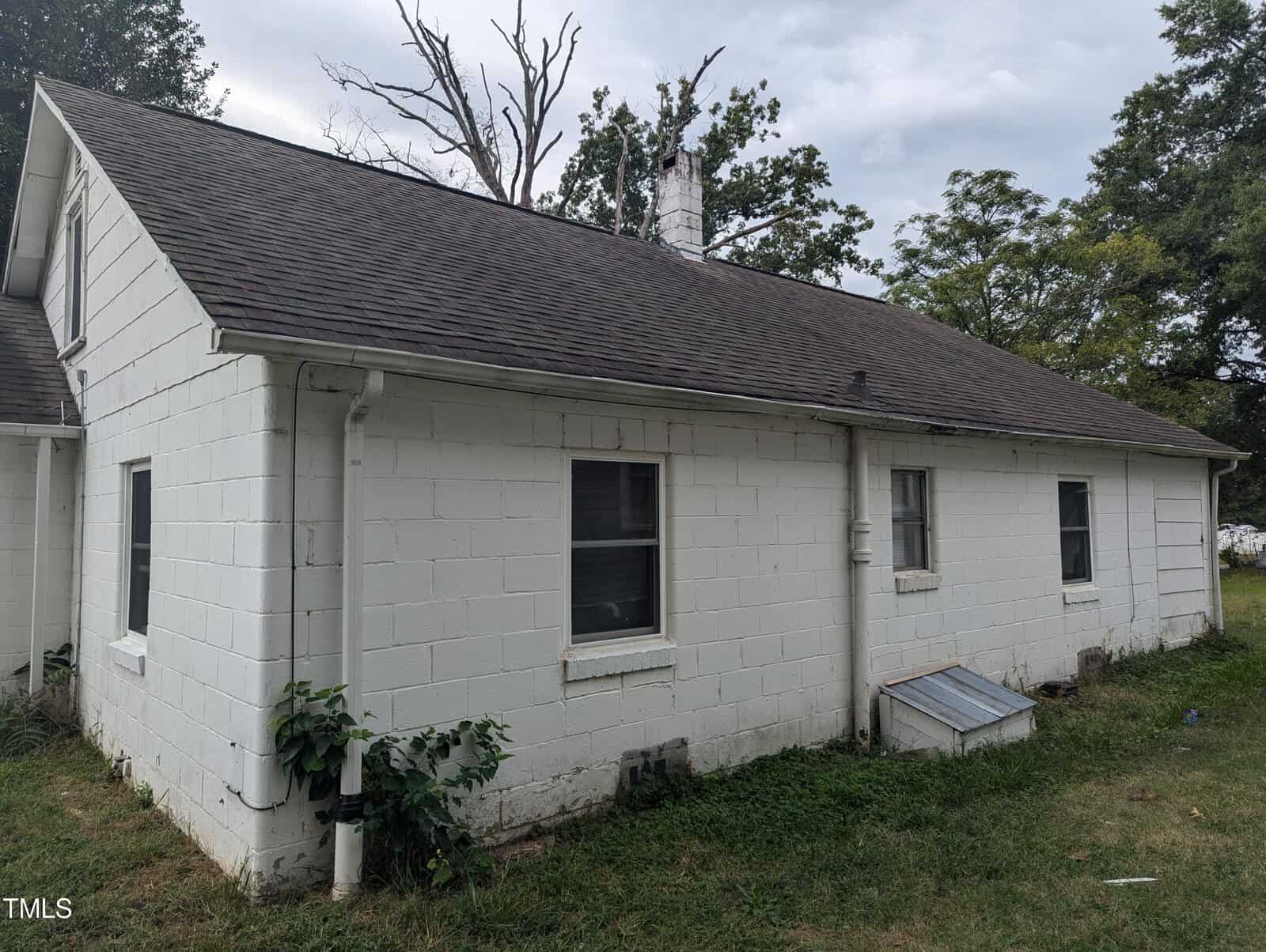 A white-painted, single-story cinder block house with a gray shingled roof. Two small rectangular windows are seen on the visible wall. The house is surrounded by overgrown grass and a few bushes. In the background, some leafless trees are standing.